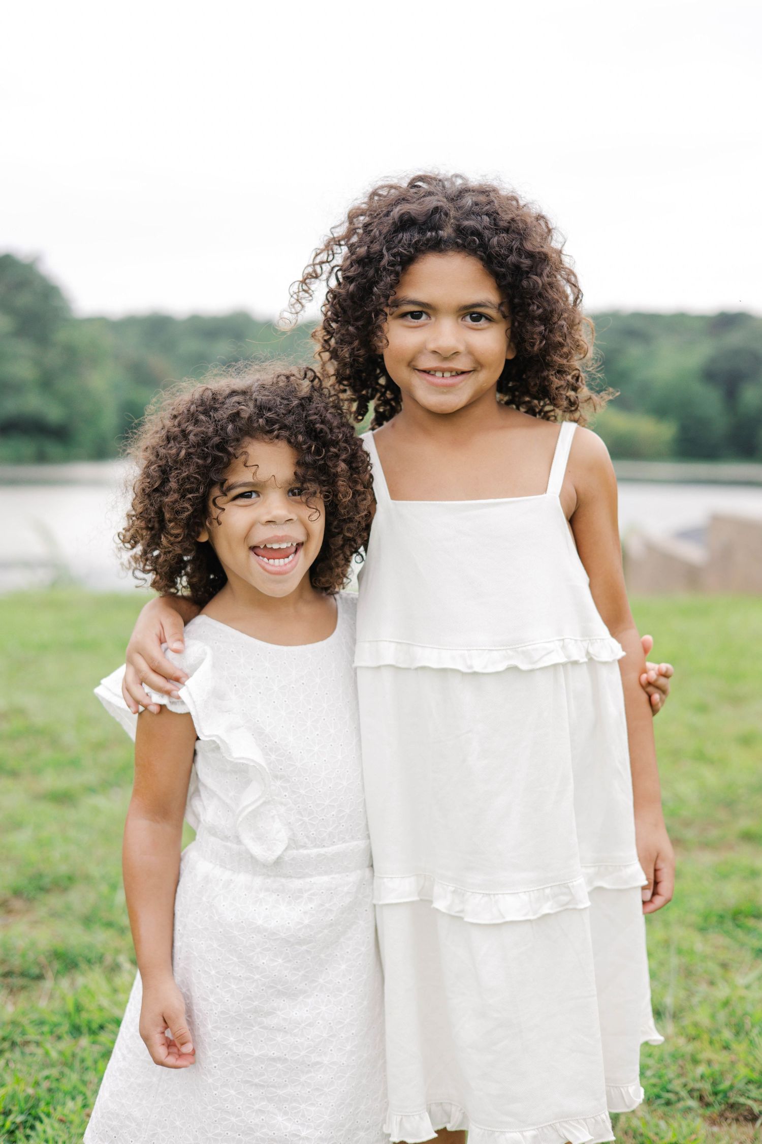 Two young girls with curly hair wearing white dresses smile together outdoors near a pond.