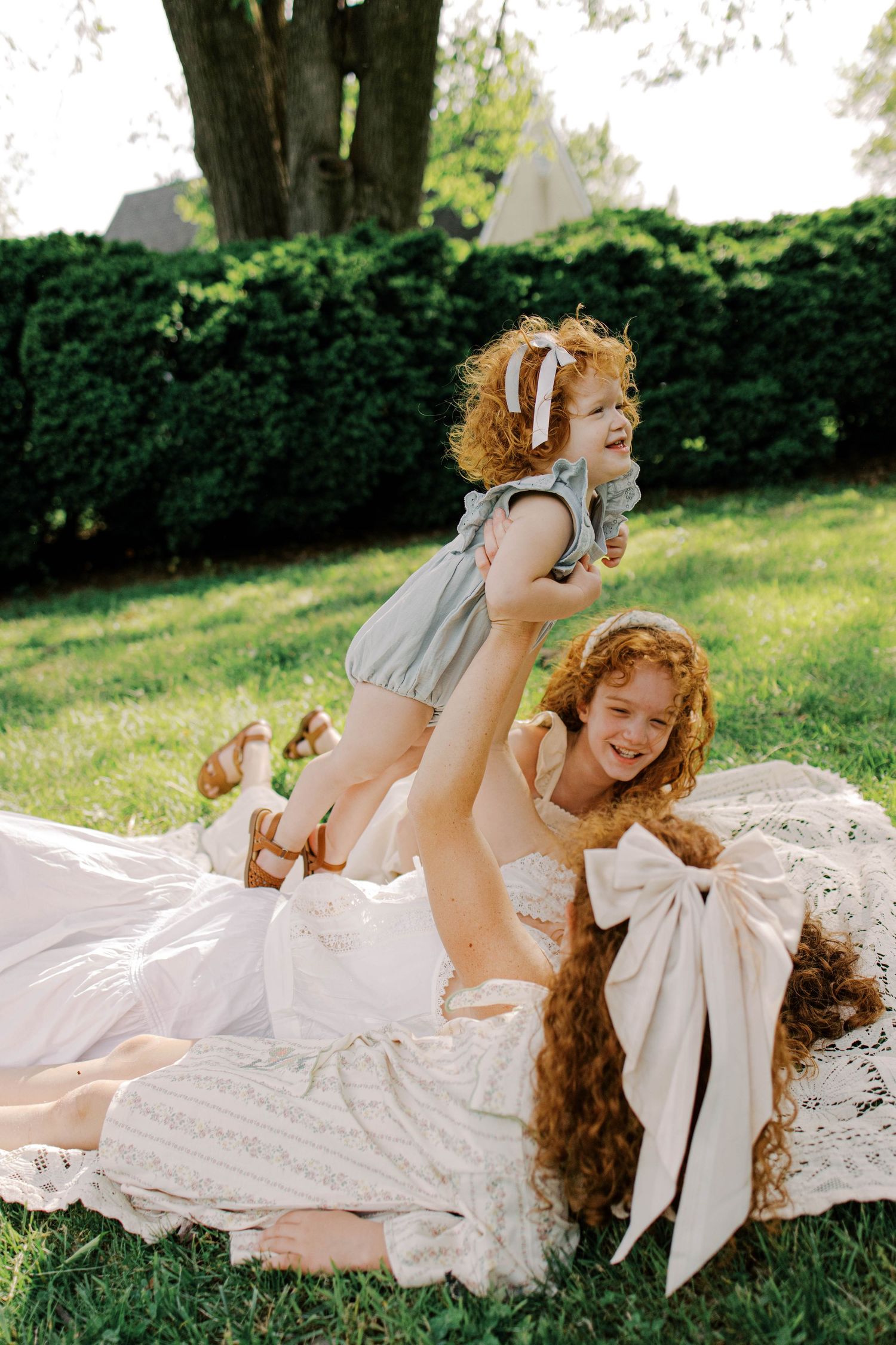 Red-haired woman in a lace dress lifts a giggling toddler overhead while another girl smiles beside them on a sunny lawn.