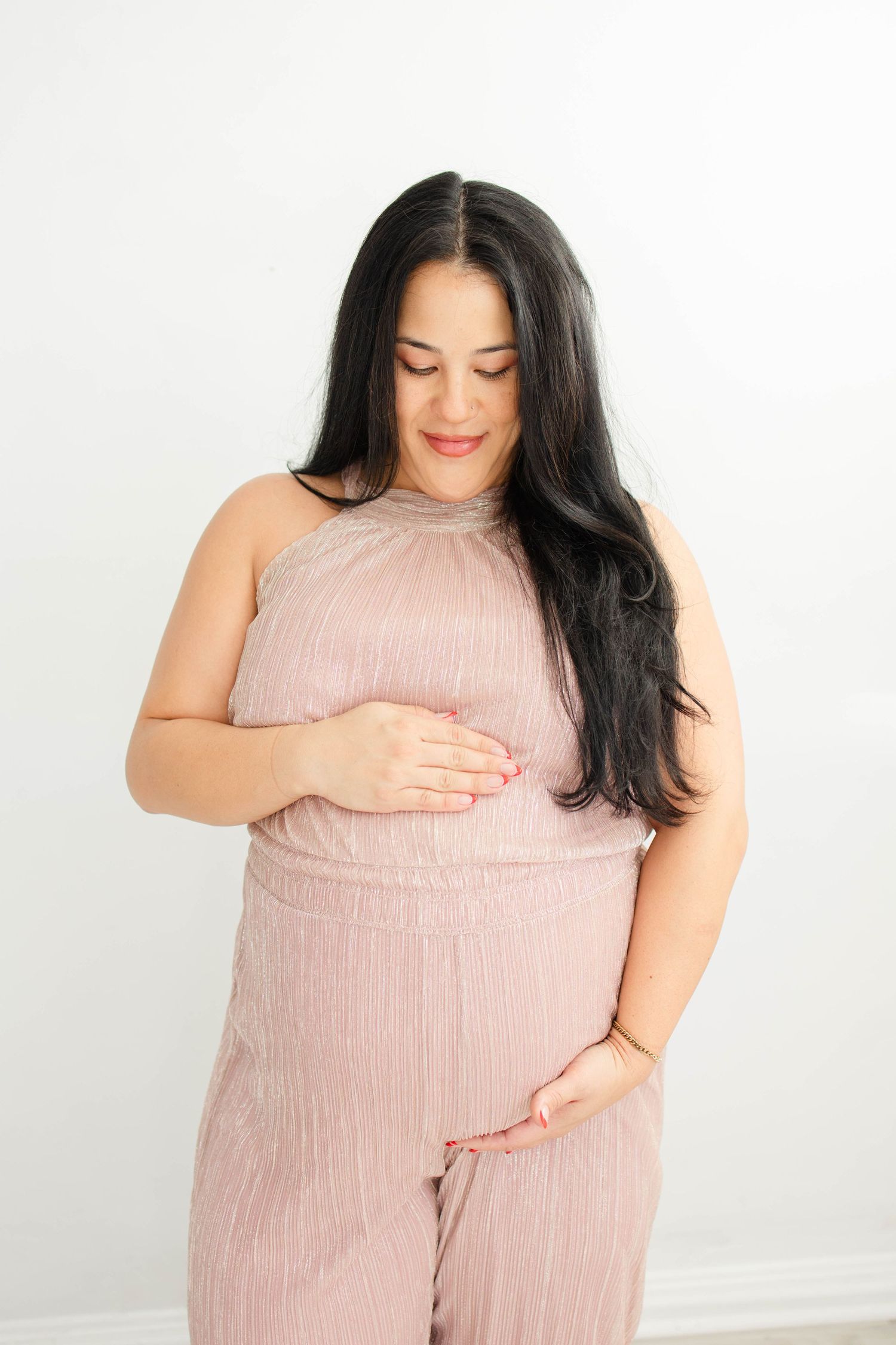 Woman in pink loungewear poses against white background during maternity photoshoot.