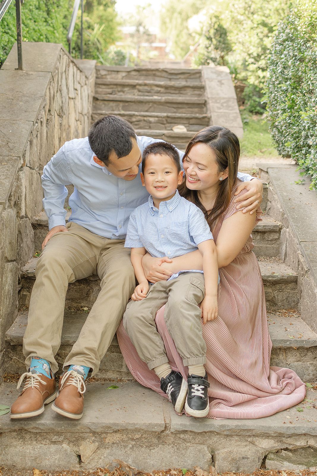 A happy Asian family of three sits on stone steps outdoors, parents smiling at their young son in matching blue shirts.