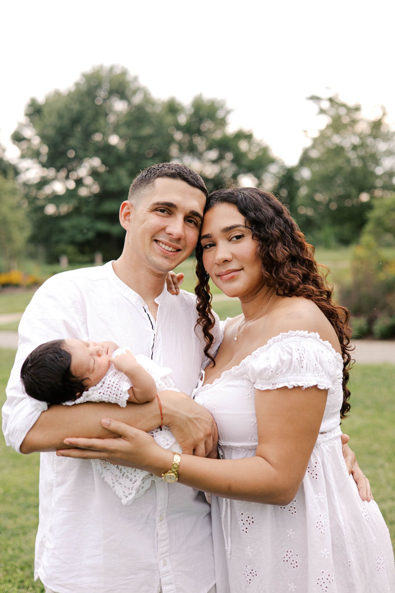 Young couple in white outfits smiling while holding their newborn baby outdoors in a lush green park.