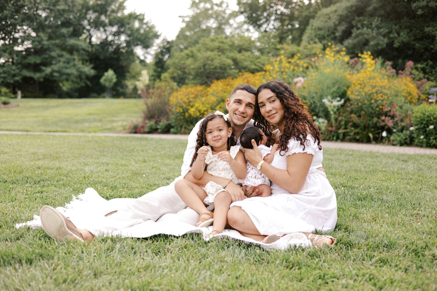 Family of four in white outfits sitting on grass in a garden park, smiling together for a portrait photo.
