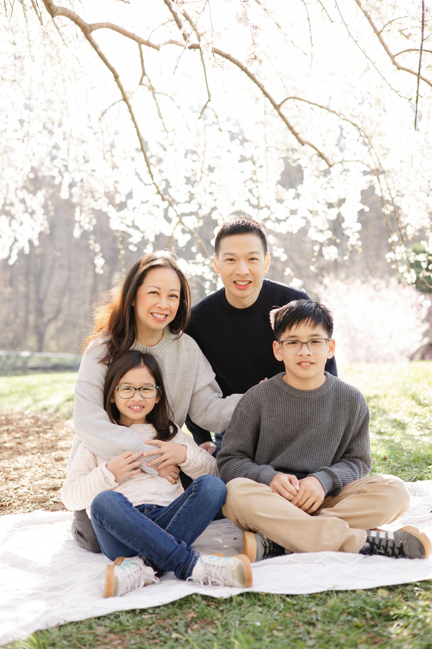 Asian family of four smiling under white cherry blossom tree, sitting on blanket outdoors in spring.