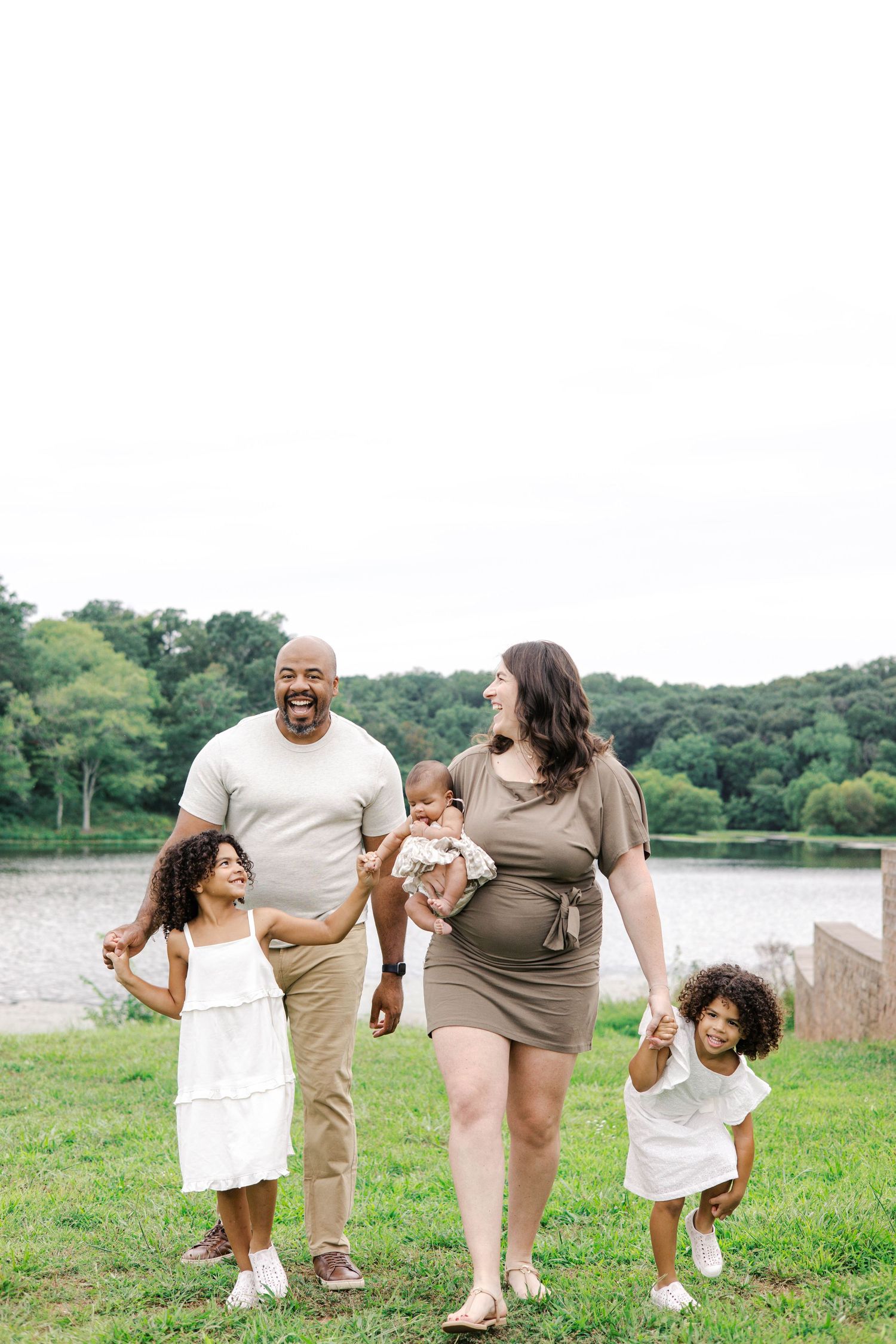 Happy interracial family walking outdoors near a lake, parents holding hands with two girls and carrying a baby.