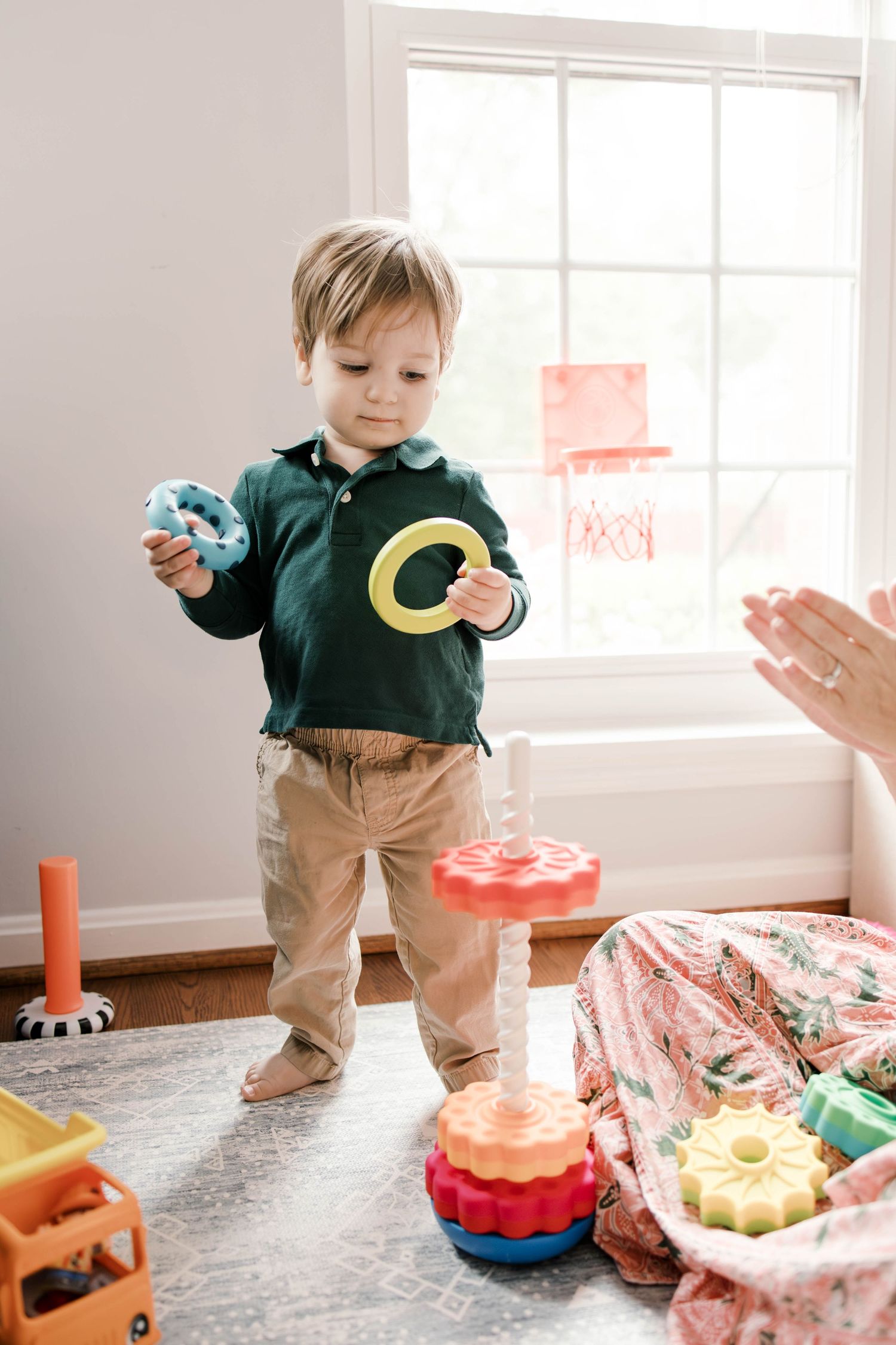 Toddler boy in green polo shirt playing with colorful stacking rings toy on carpet in bright playroom.