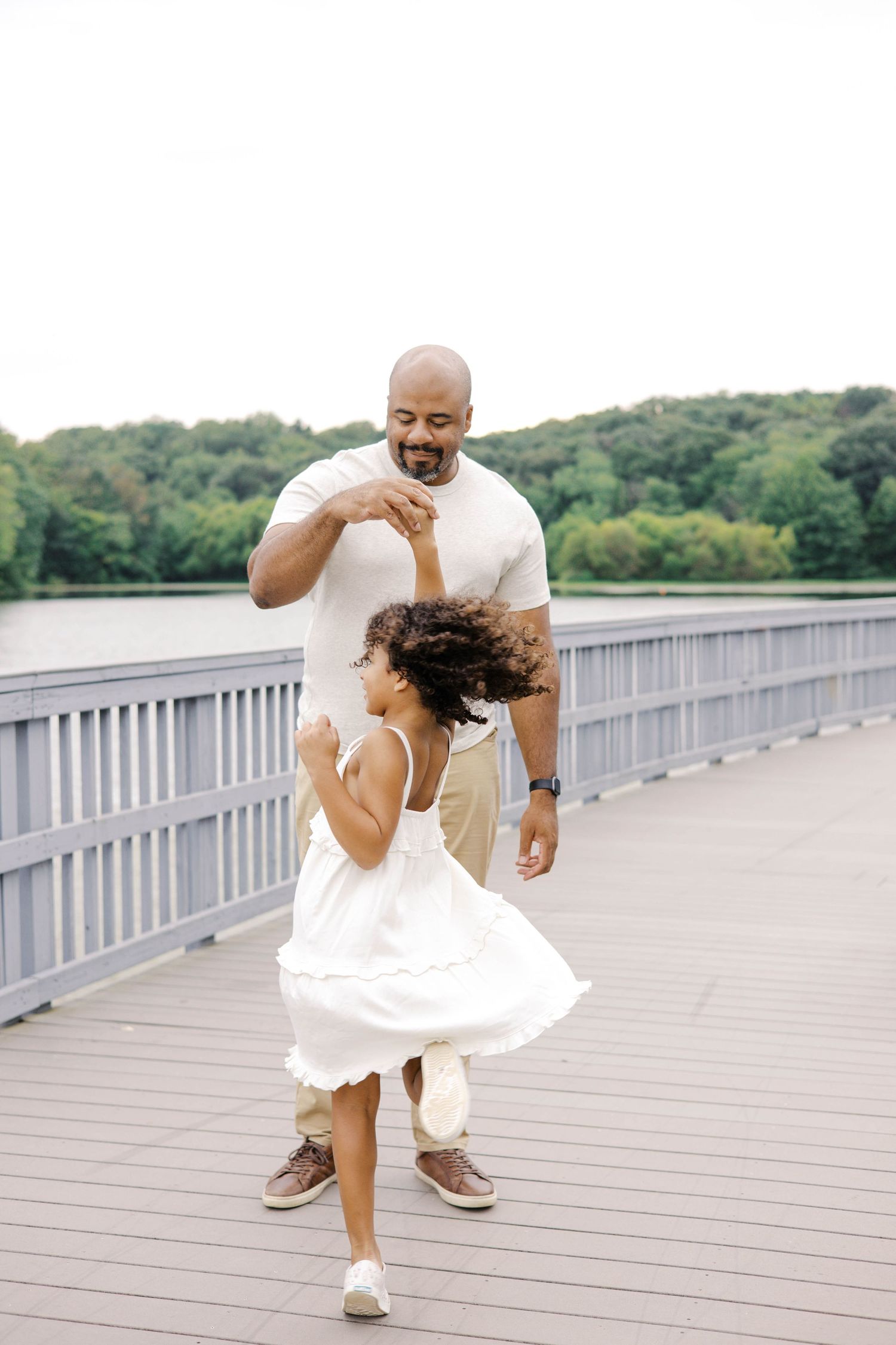 A father and daughter dancing joyfully on a wooden bridge near a lake surrounded by lush green trees.
