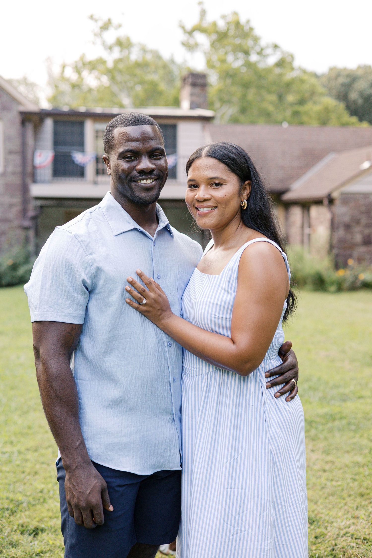 A smiling couple poses outdoors in coordinating blue outfits in front of a brick house surrounded by green trees.