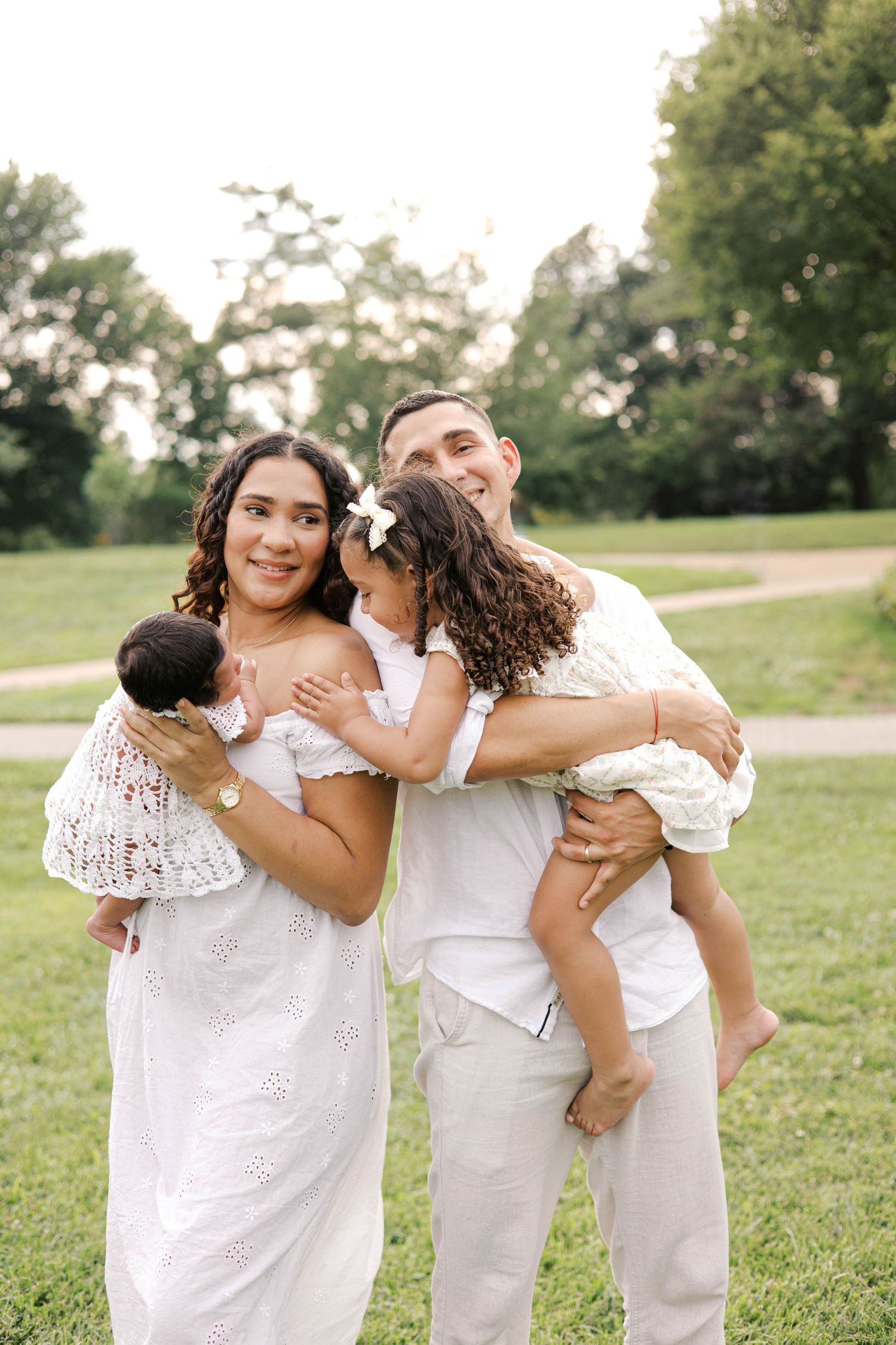 Happy family in white outfits posing outdoors, parents holding two young children in a lush green park setting.