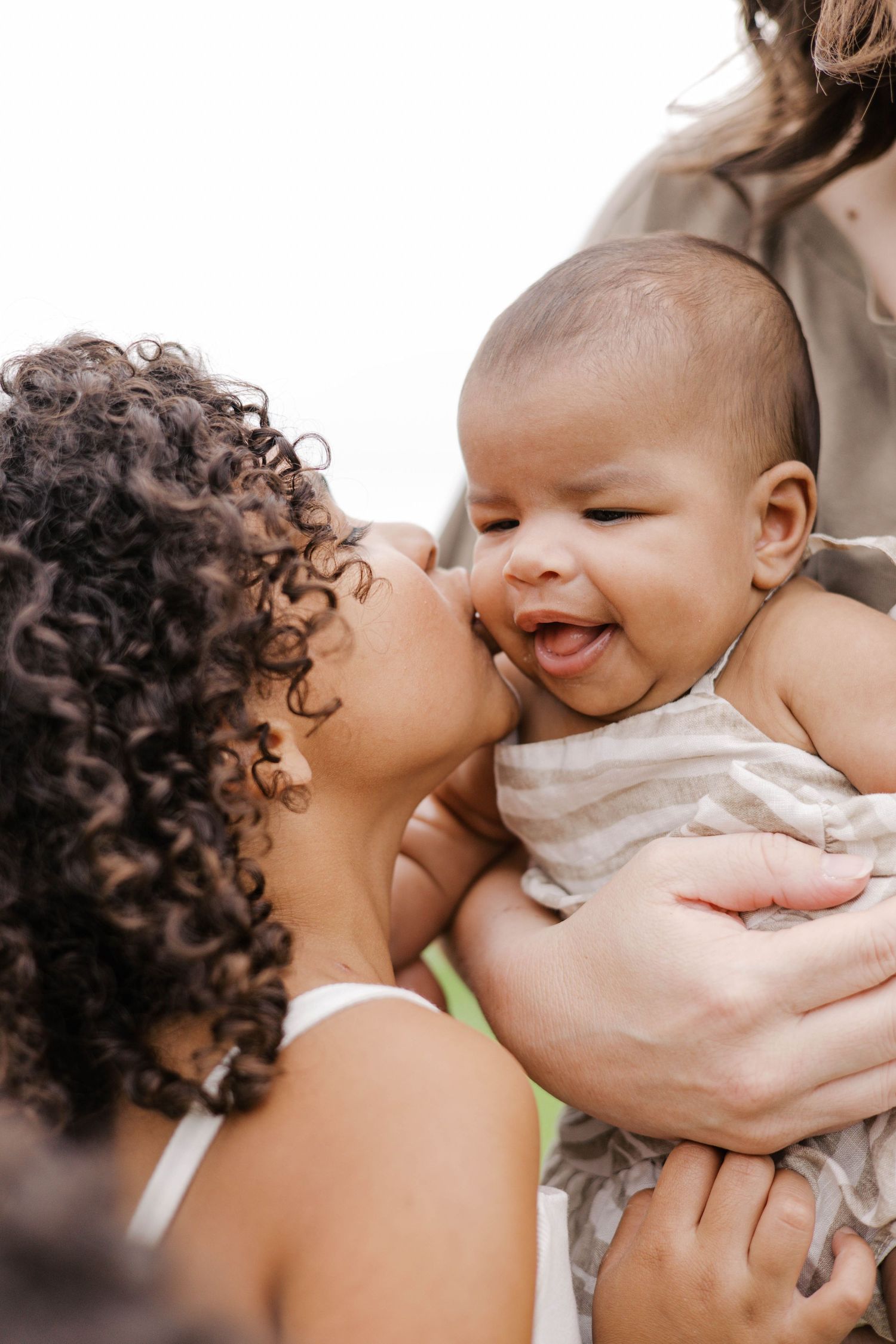A curly-haired woman kisses a laughing baby being held by another person outdoors.