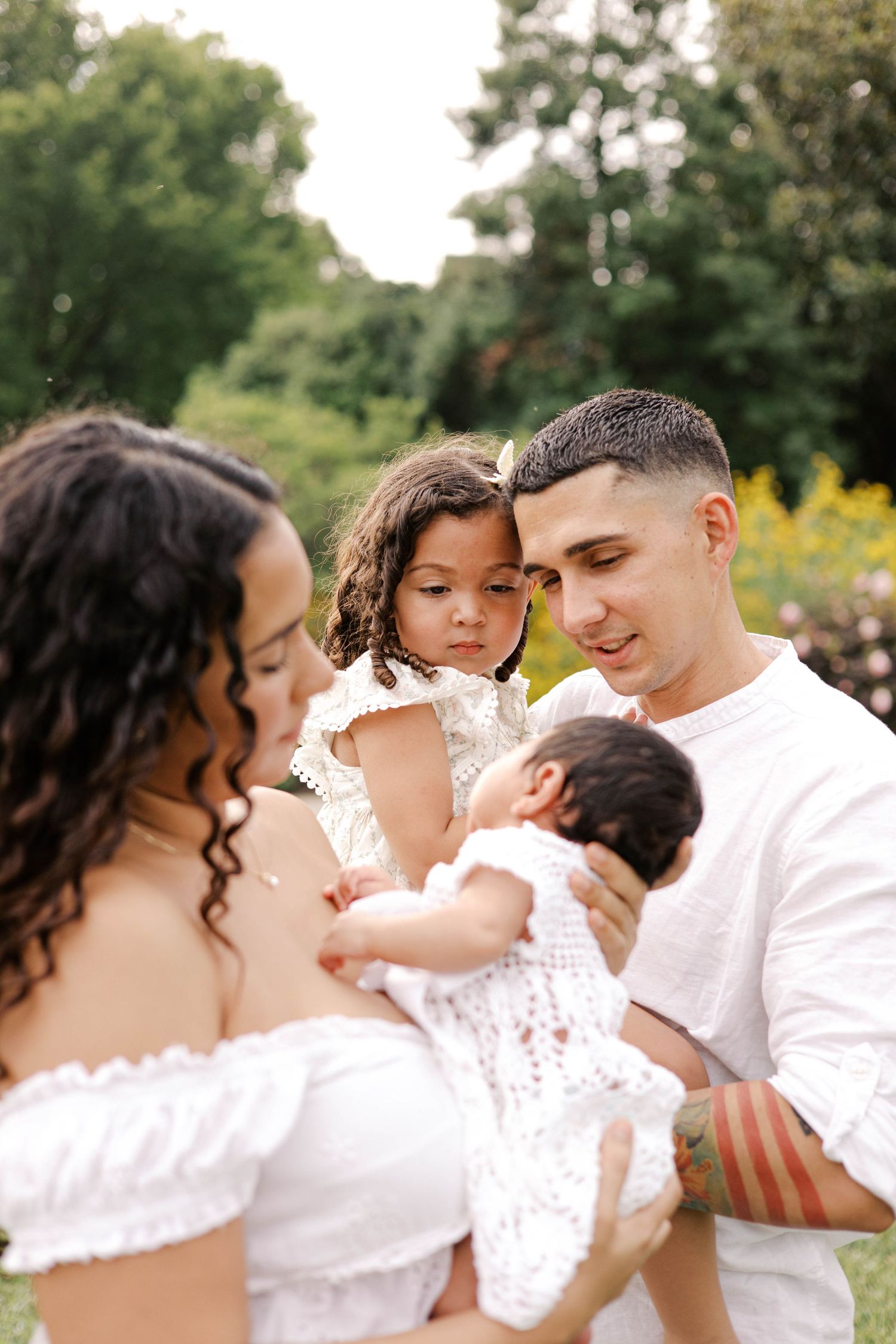 Family in white outfits outdoors, parents lovingly gazing at newborn baby while young daughter looks on.