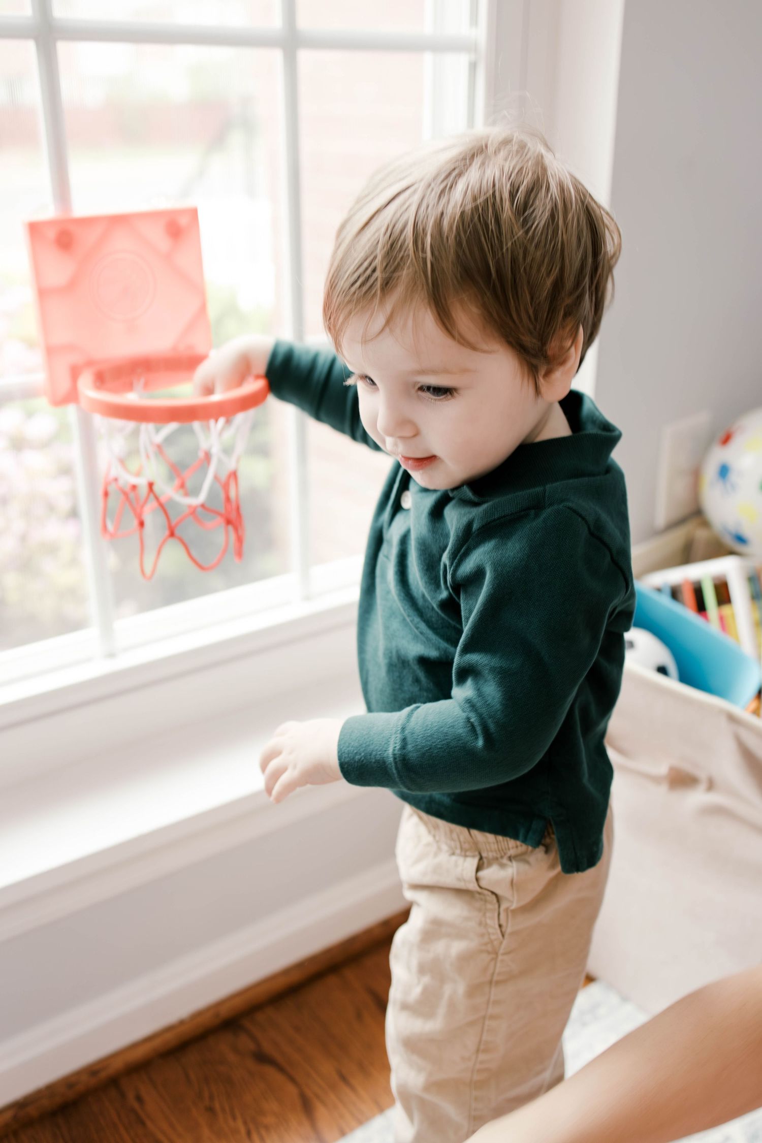 Toddler boy in green shirt and khaki pants playing with a mini basketball hoop by a bright window indoors.