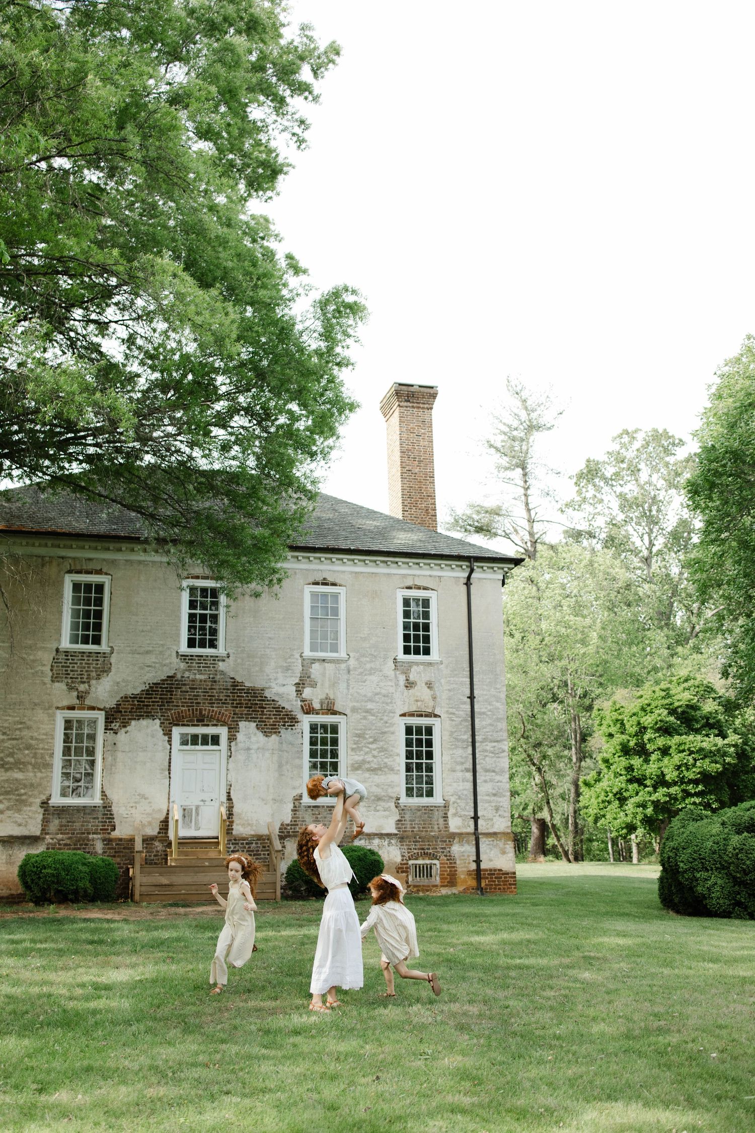 Mother in a white dress lifts a child in the air as two girls play on the lawn in front of a historic brick manor house.