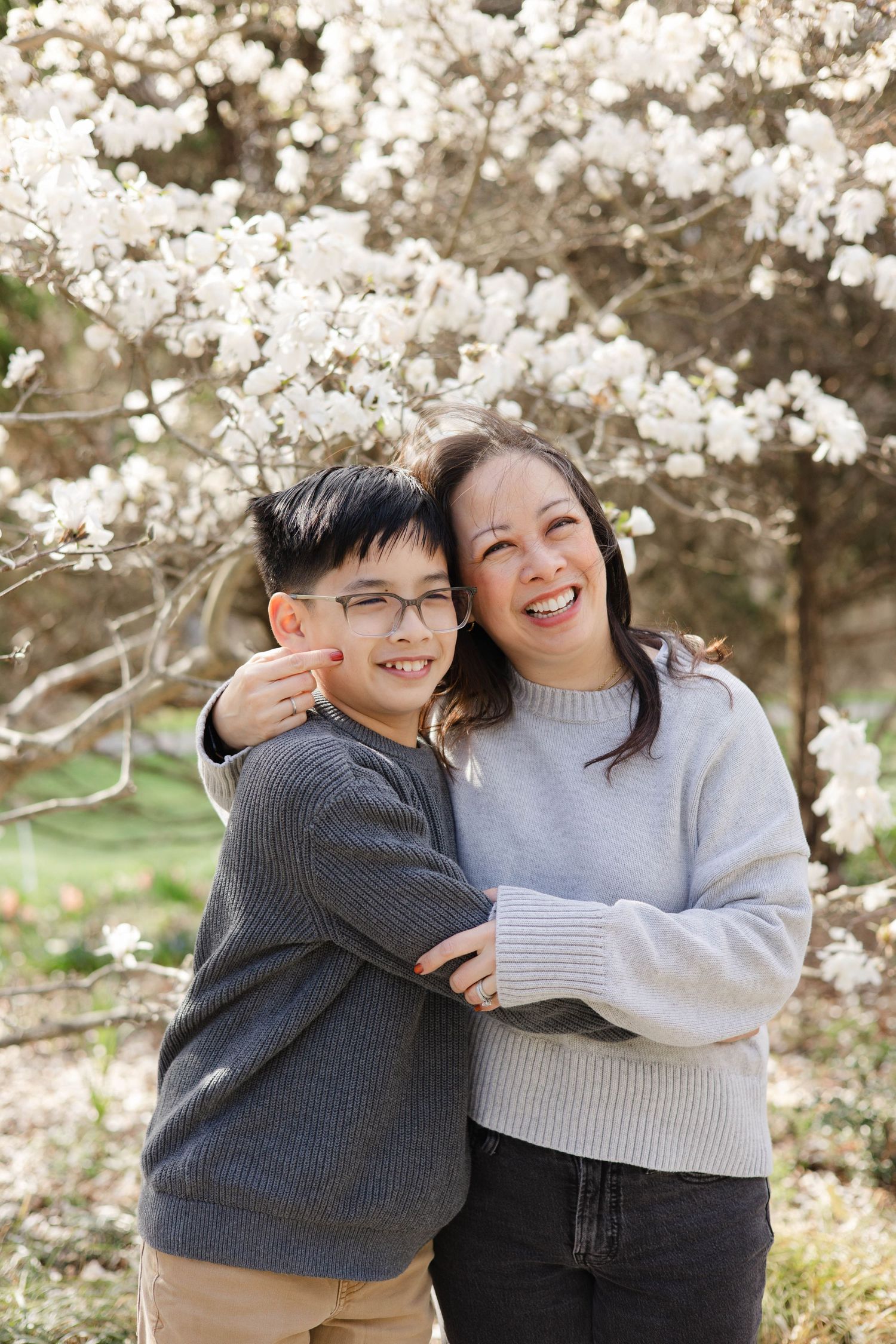 A smiling woman and young boy with glasses embrace outdoors near a blooming white magnolia tree in spring.