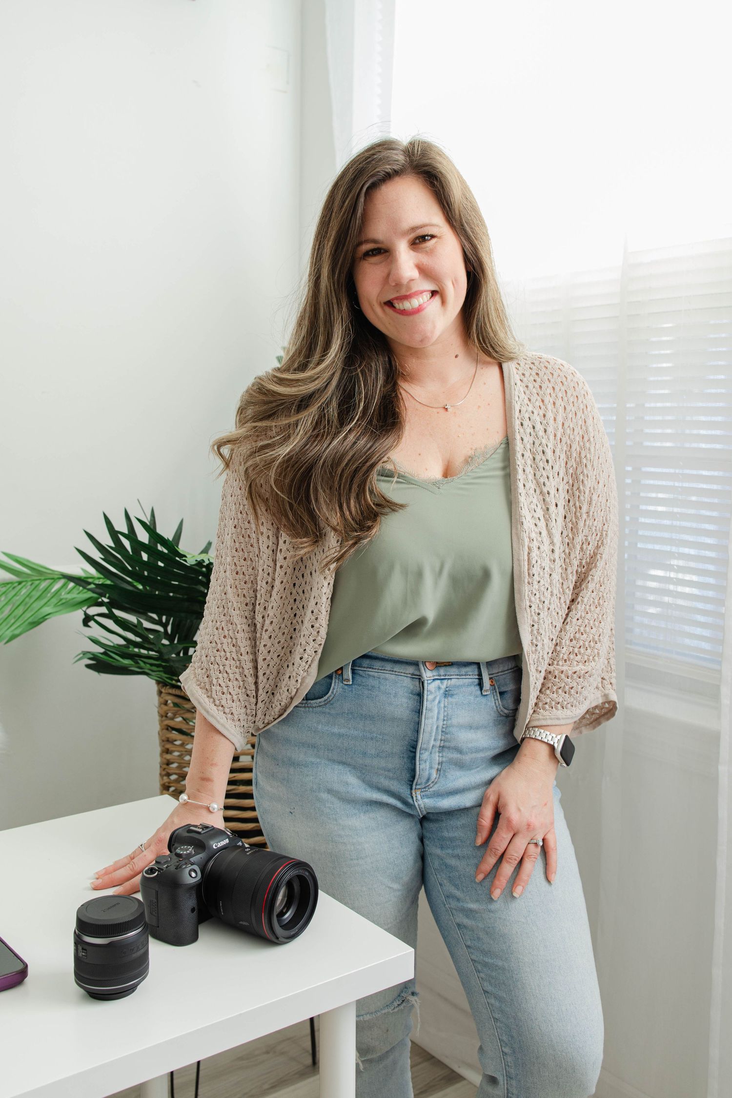 Photographer in beige cardigan and green top sits at white desk with camera equipment and plant decor.