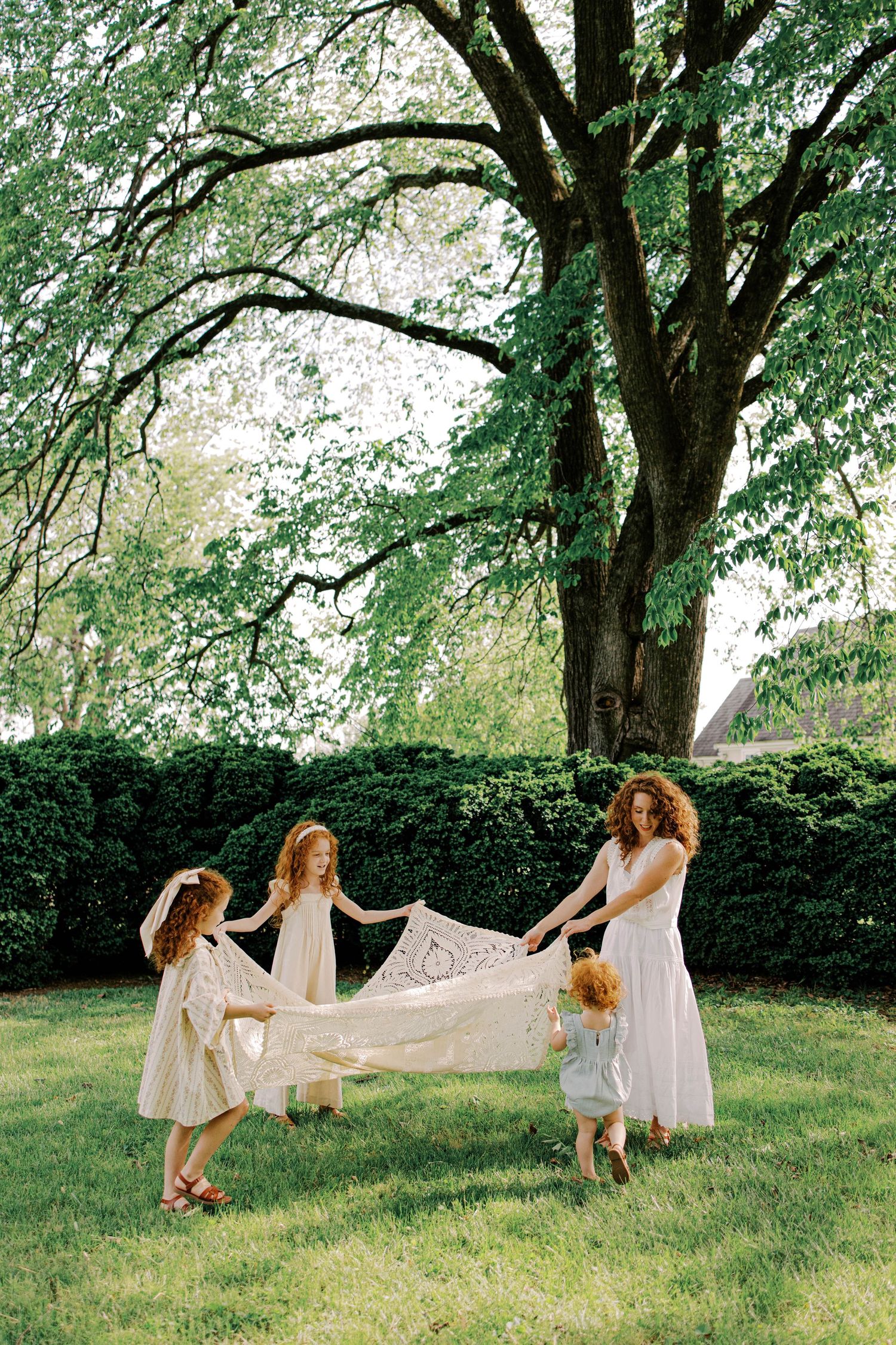 A woman and three young girls in light dresses hold a lace blanket on a sunny lawn beneath a large tree.