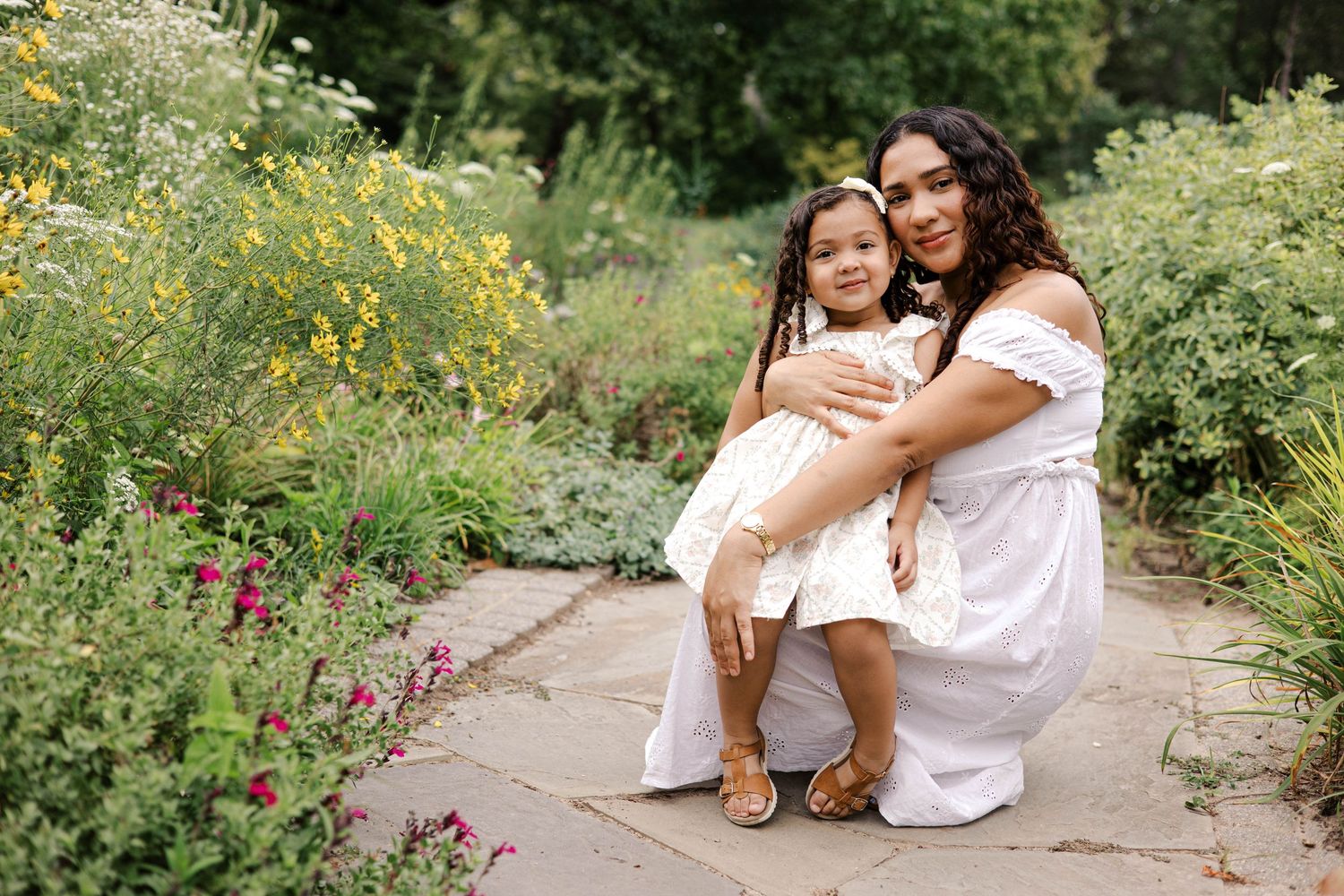 A mother and young daughter in matching white dresses pose together in a lush garden with colorful flowers.