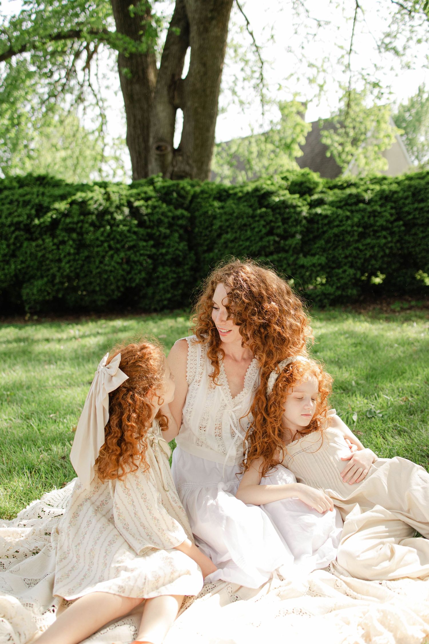 A curly redhead mother sits on a blanket outdoors with her two redheaded daughters in white dresses.