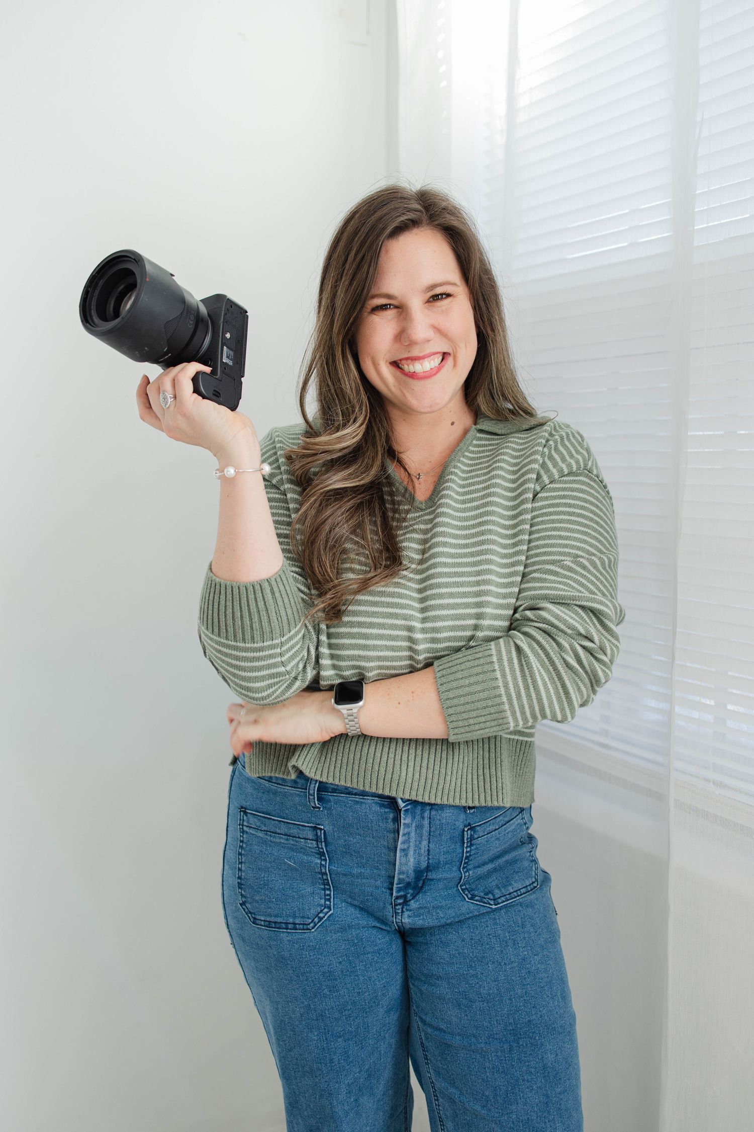 Photographer in green striped shirt and jeans demonstrates camera poses against white wall.