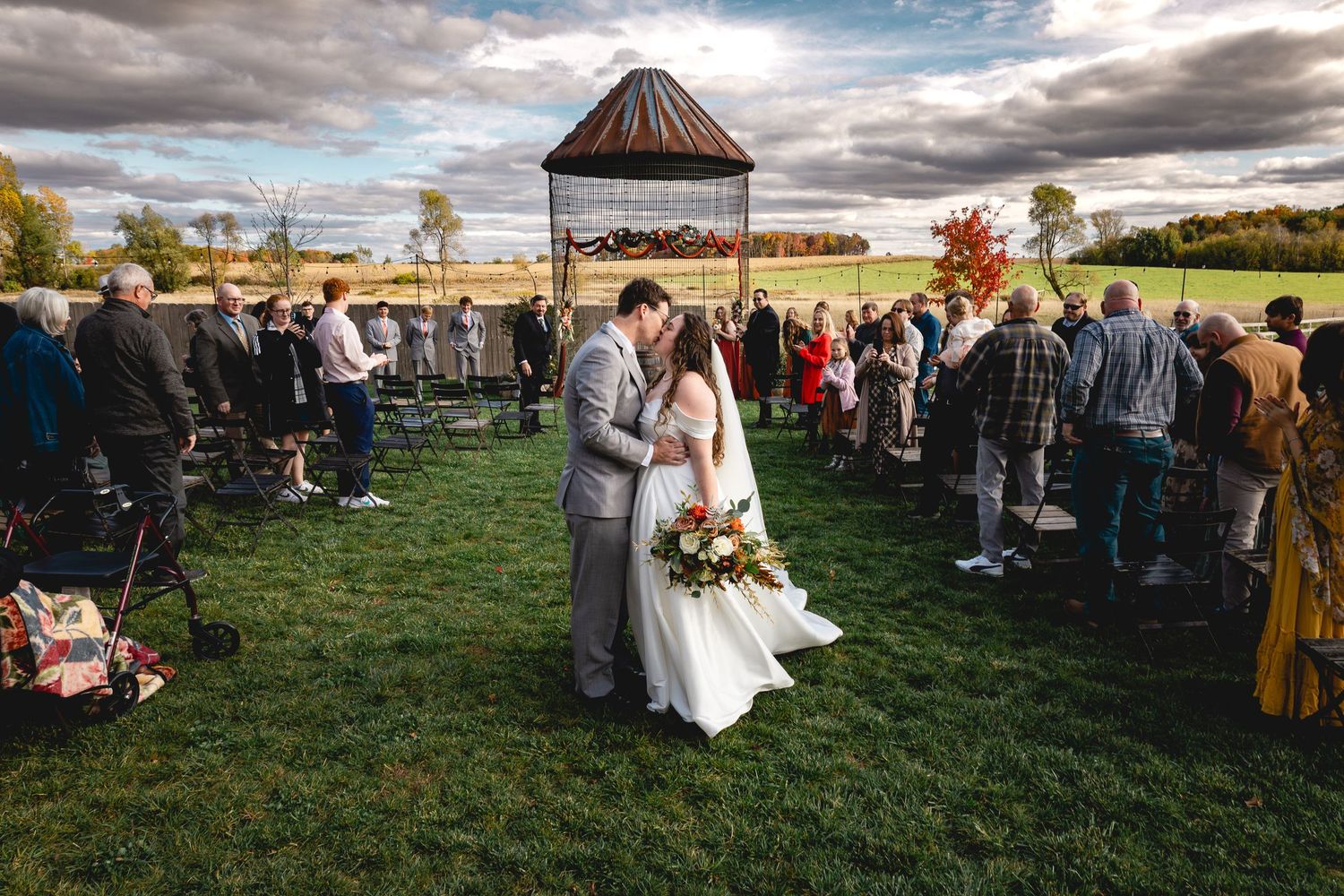 Outdoor wedding ceremony gathering with guests on grassy field under cloudy skies.