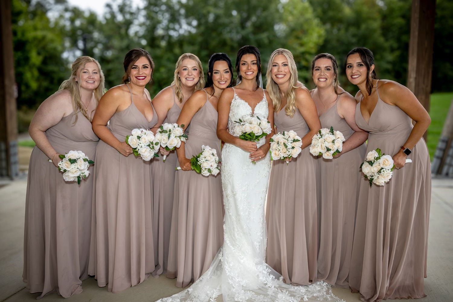 Bridesmaids in taupe dresses hold white bouquets while posing together outdoors.