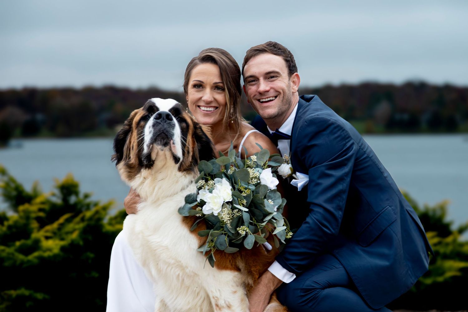 A joyful wedding couple embraces outdoors with a lake and autumn trees in the background.