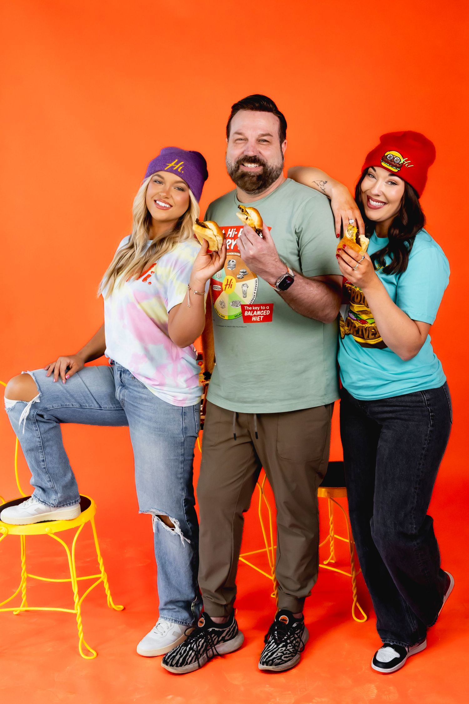 Three friends pose with snacks against bright orange background in casual attire.
