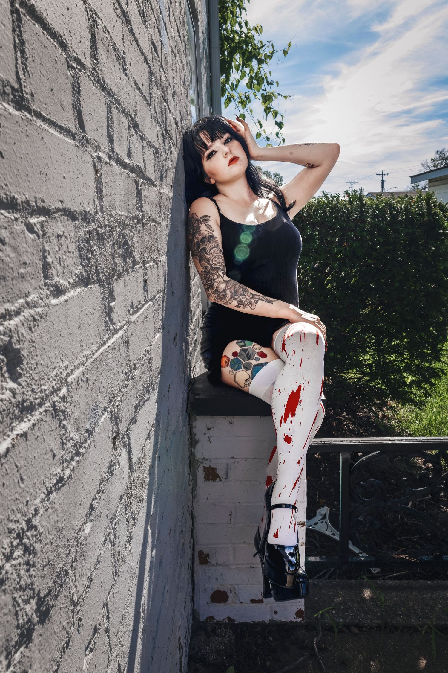 Model in black top with tattoos poses against white brick wall outdoors.