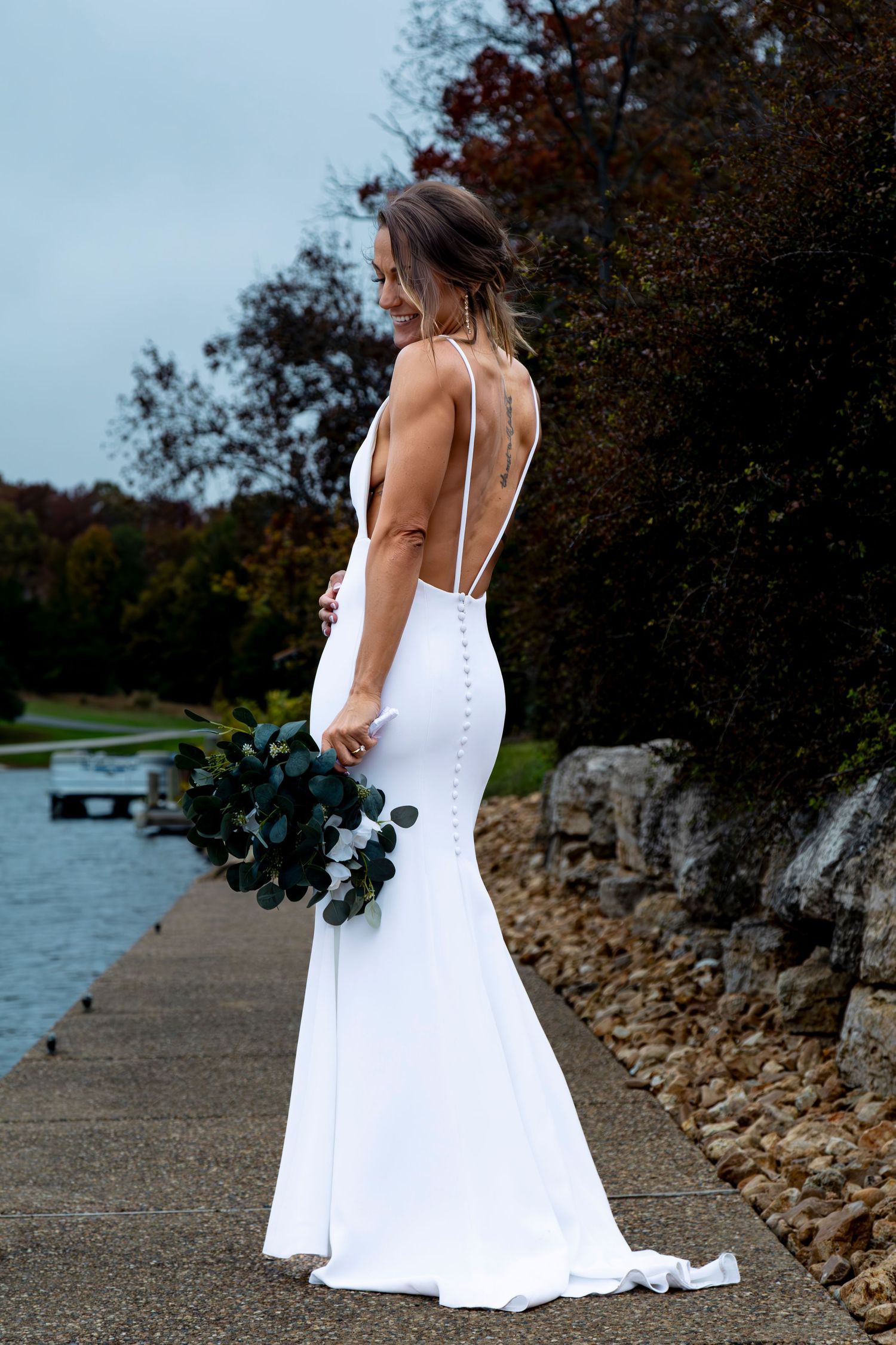 A bride in a white wedding dress poses along a waterfront path with stone barriers.