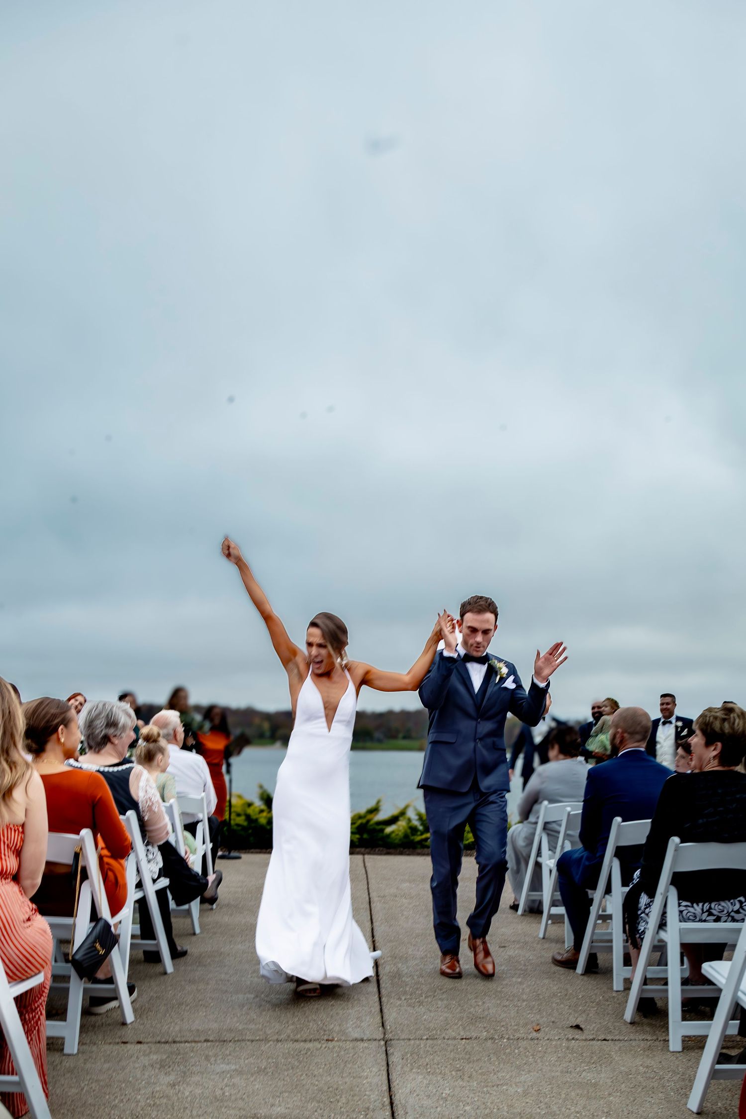 Wedding guests seated in white chairs at an outdoor waterfront ceremony react with joy.