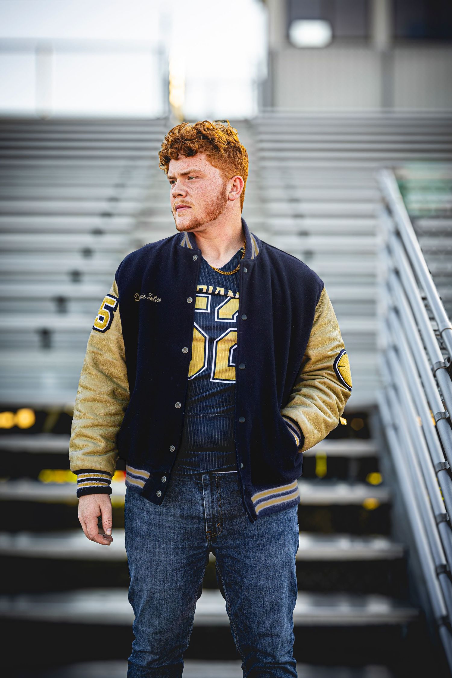 Person wearing varsity jacket and jeans poses near stadium seating.