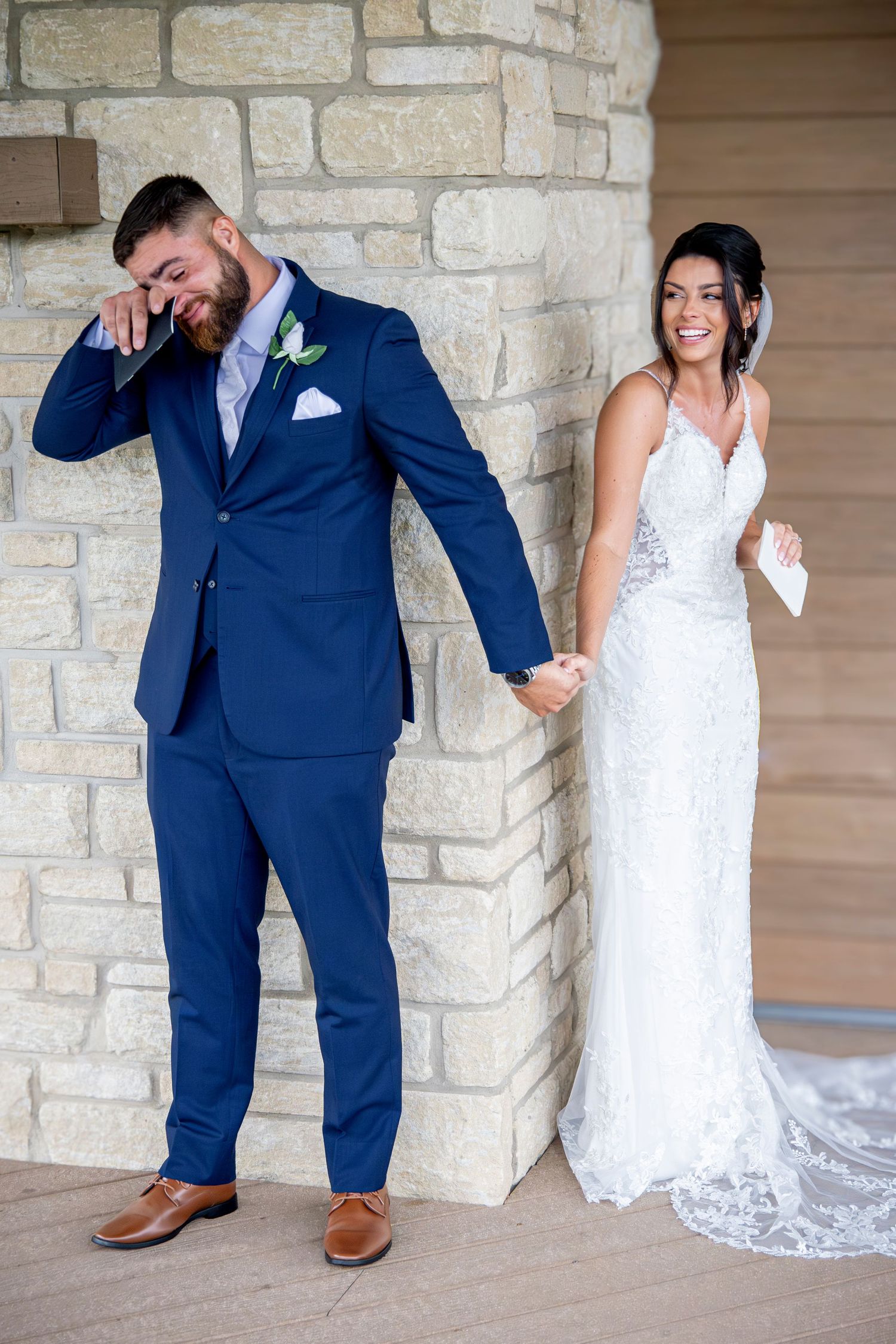 Playful wedding photos taken against a stone wall backdrop with navy suit and white lace dress.