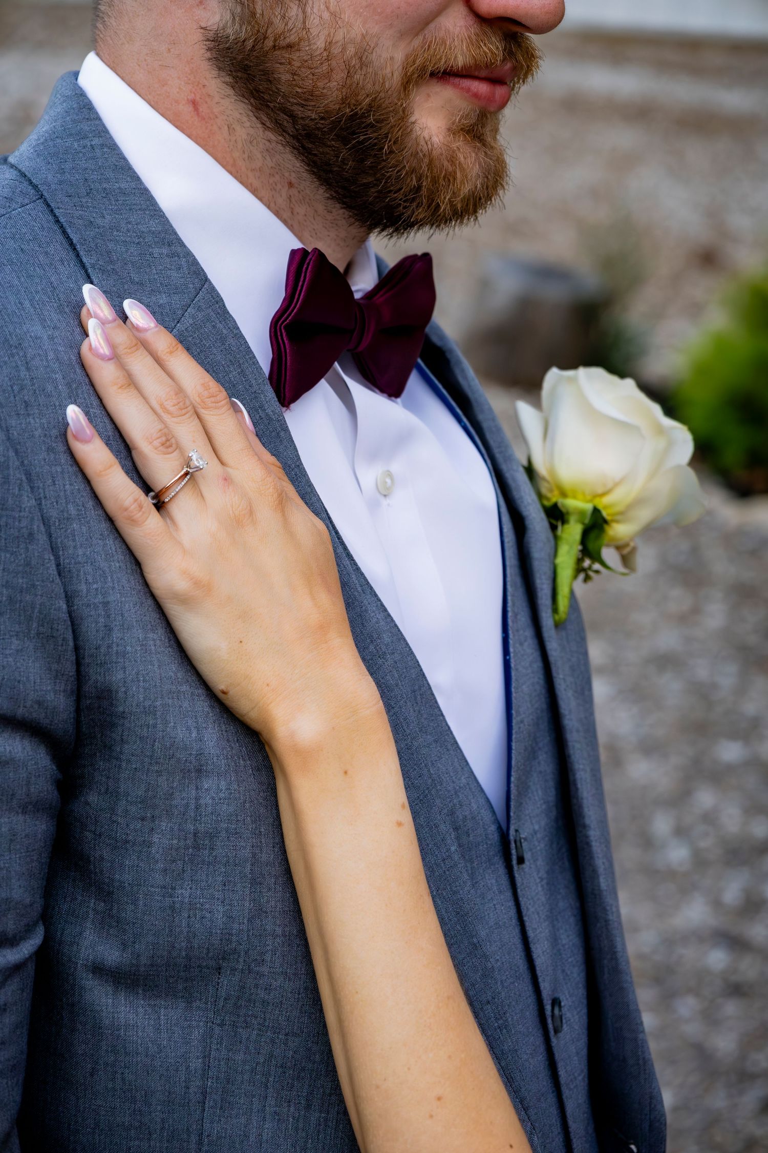 Close-up details of wedding ring and boutonniere with hands against grey suit.