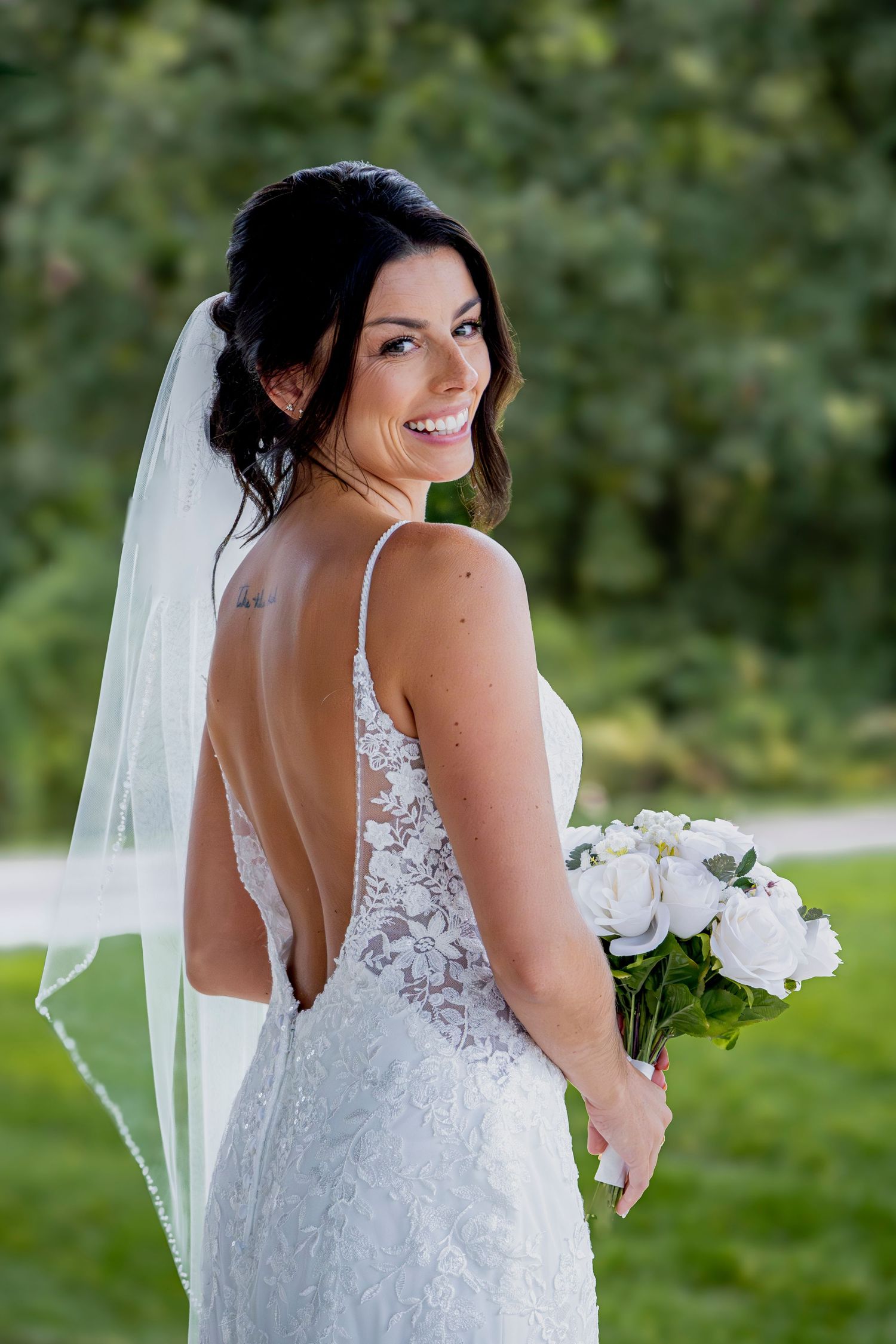 Bride in detailed lace gown poses with white bouquet in garden setting.