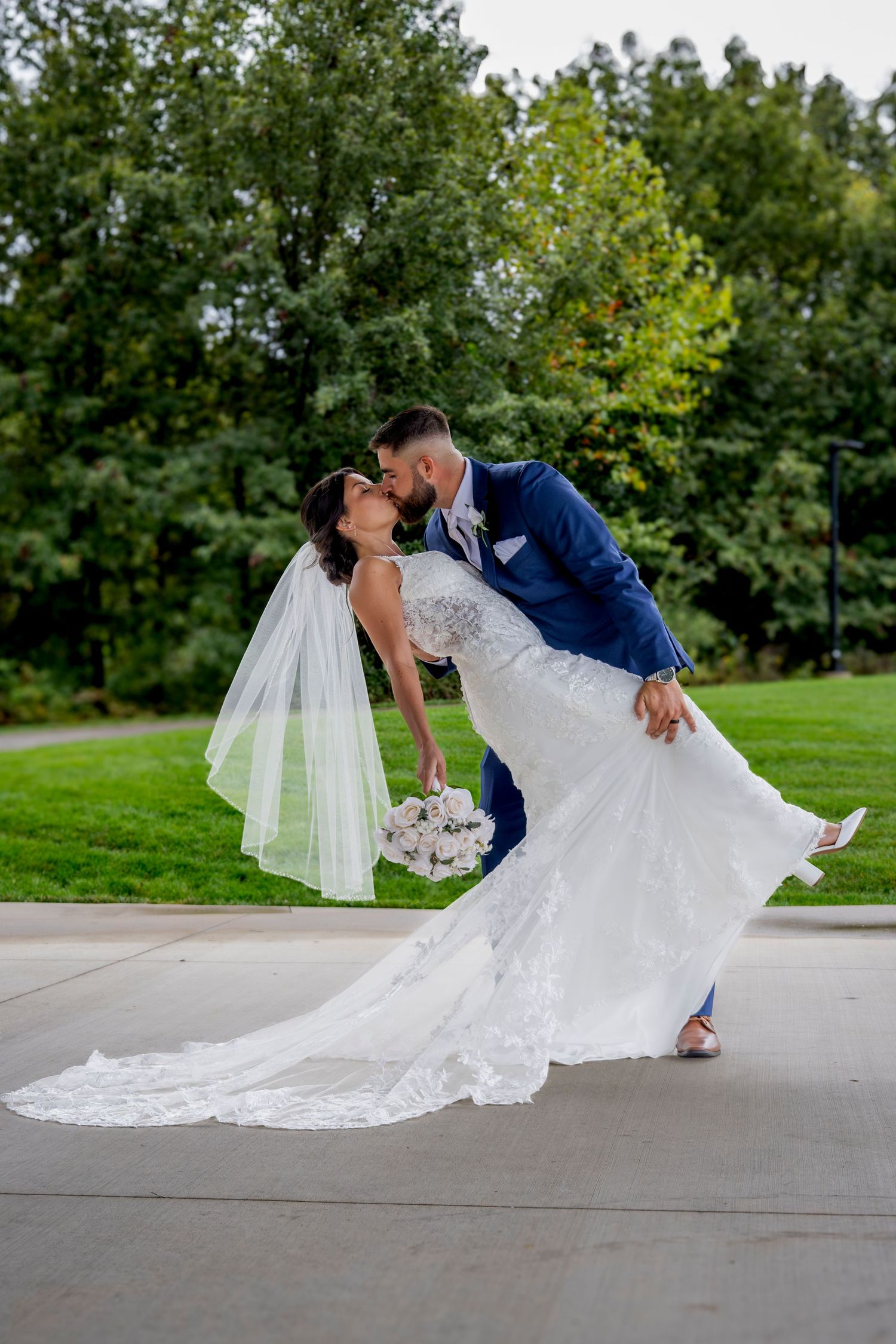 Dramatic dip pose wedding photos captured on concrete path with green foliage background.