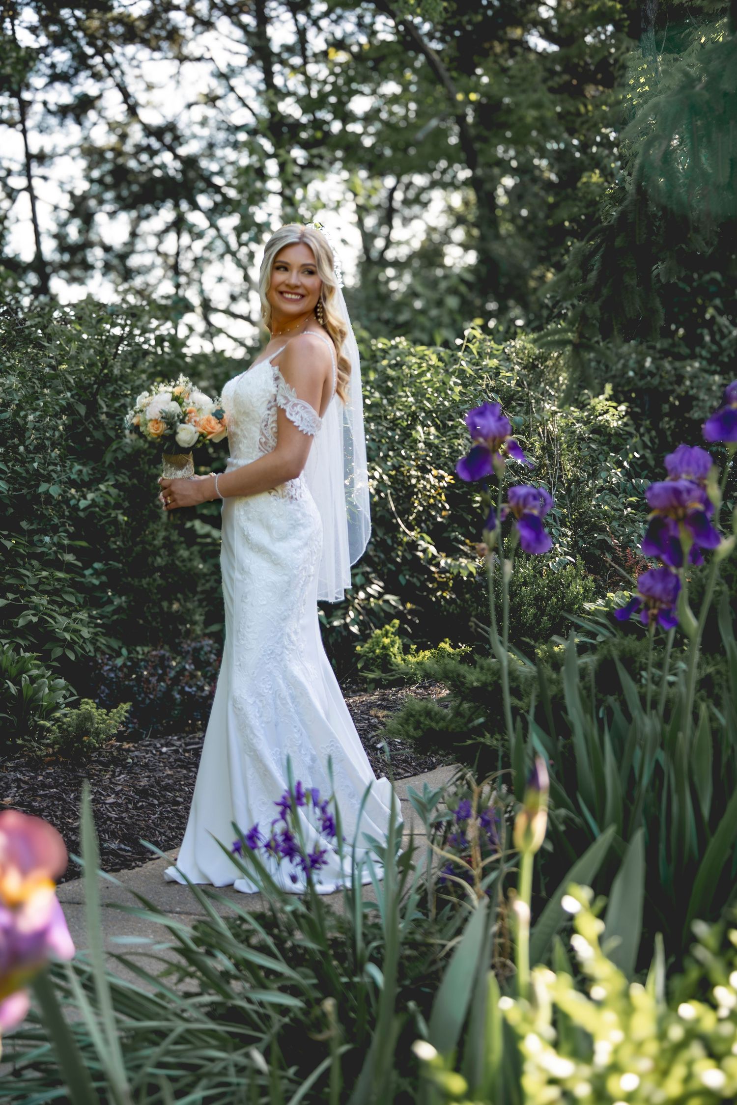 A bride poses in a garden surrounded by blooming purple iris flowers and greenery.