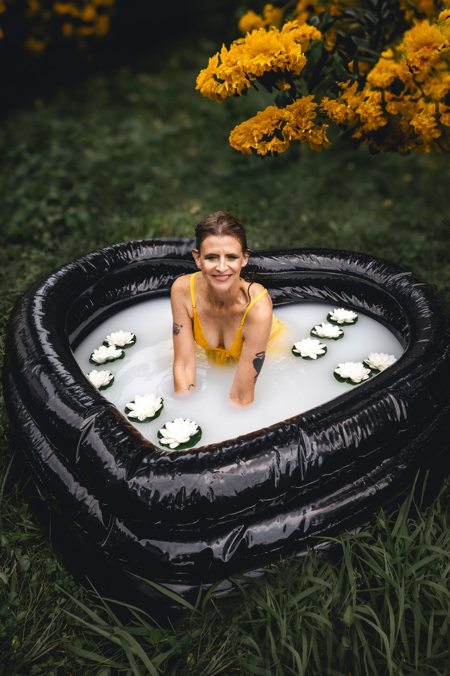 Person relaxing in heart-shaped pool with floating water lilies and yellow flowers overhead.