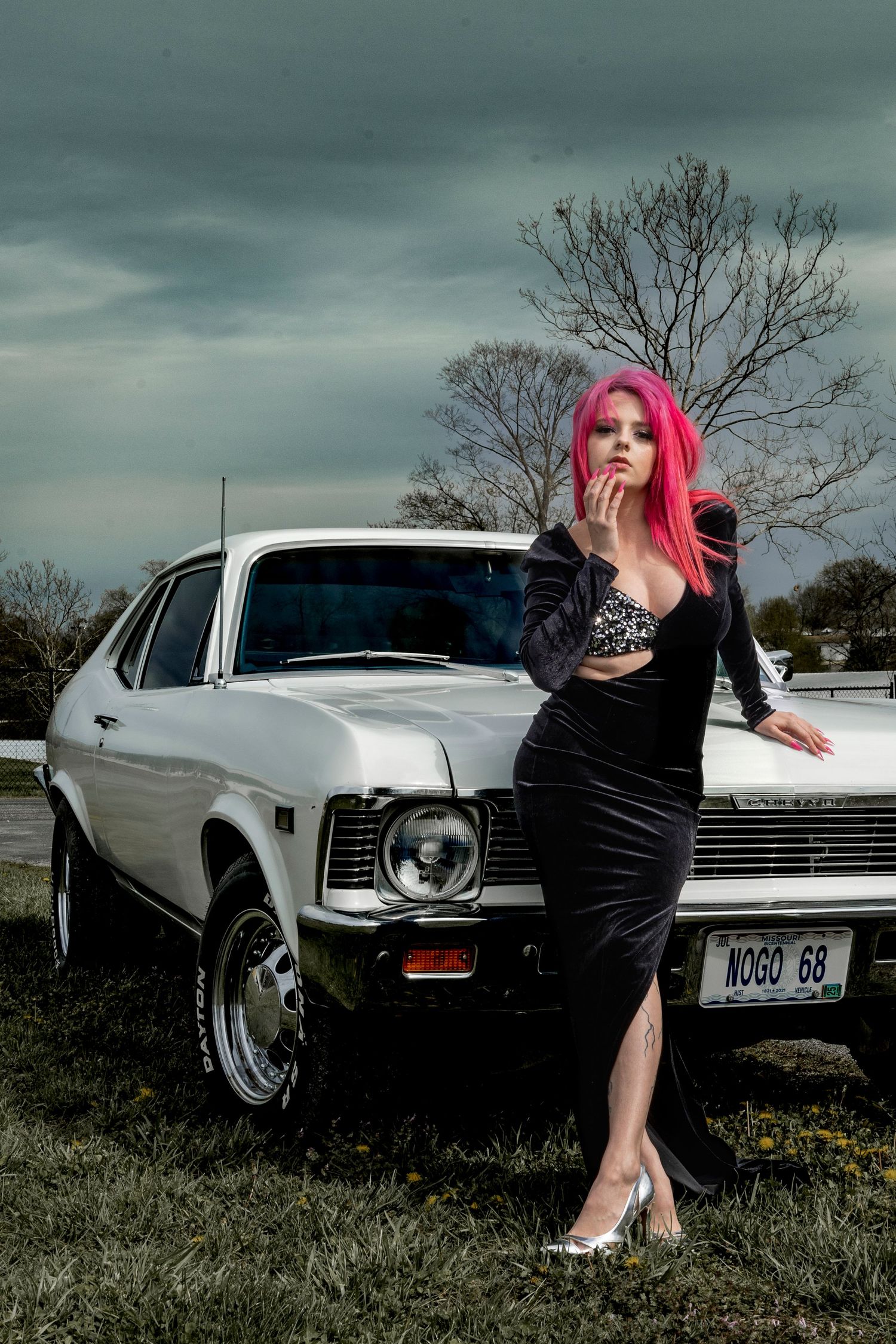 Fashion shoot featuring pink-haired model in black dress leaning against classic white Chevy Nova.