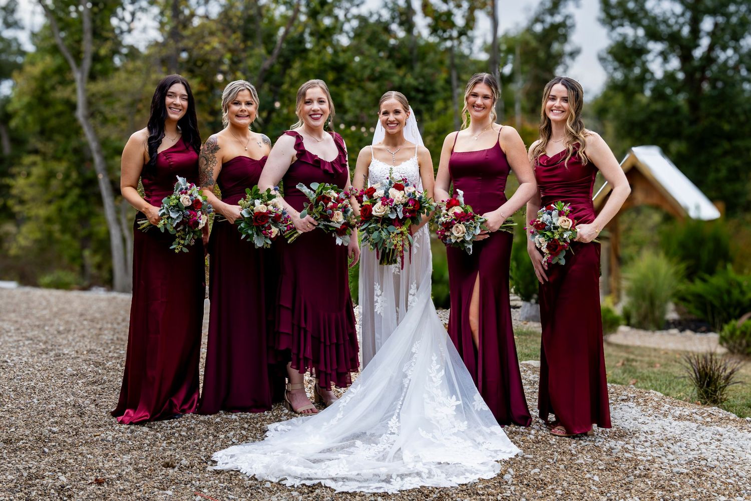 Wedding party in burgundy dresses poses outdoors on gravel path.