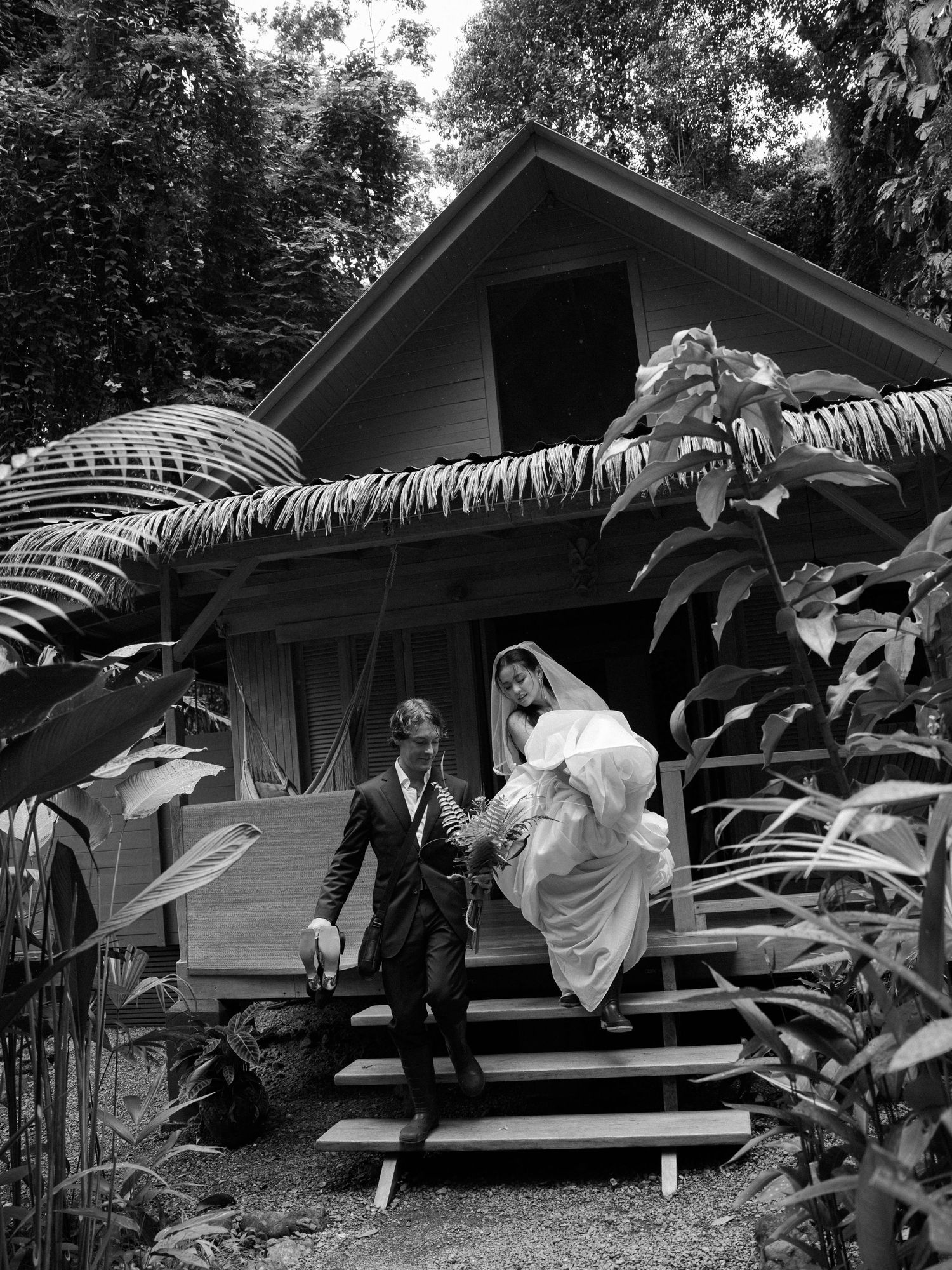 Couple in wedding attire descending stairs of tropical cottage surrounded by lush foliage.