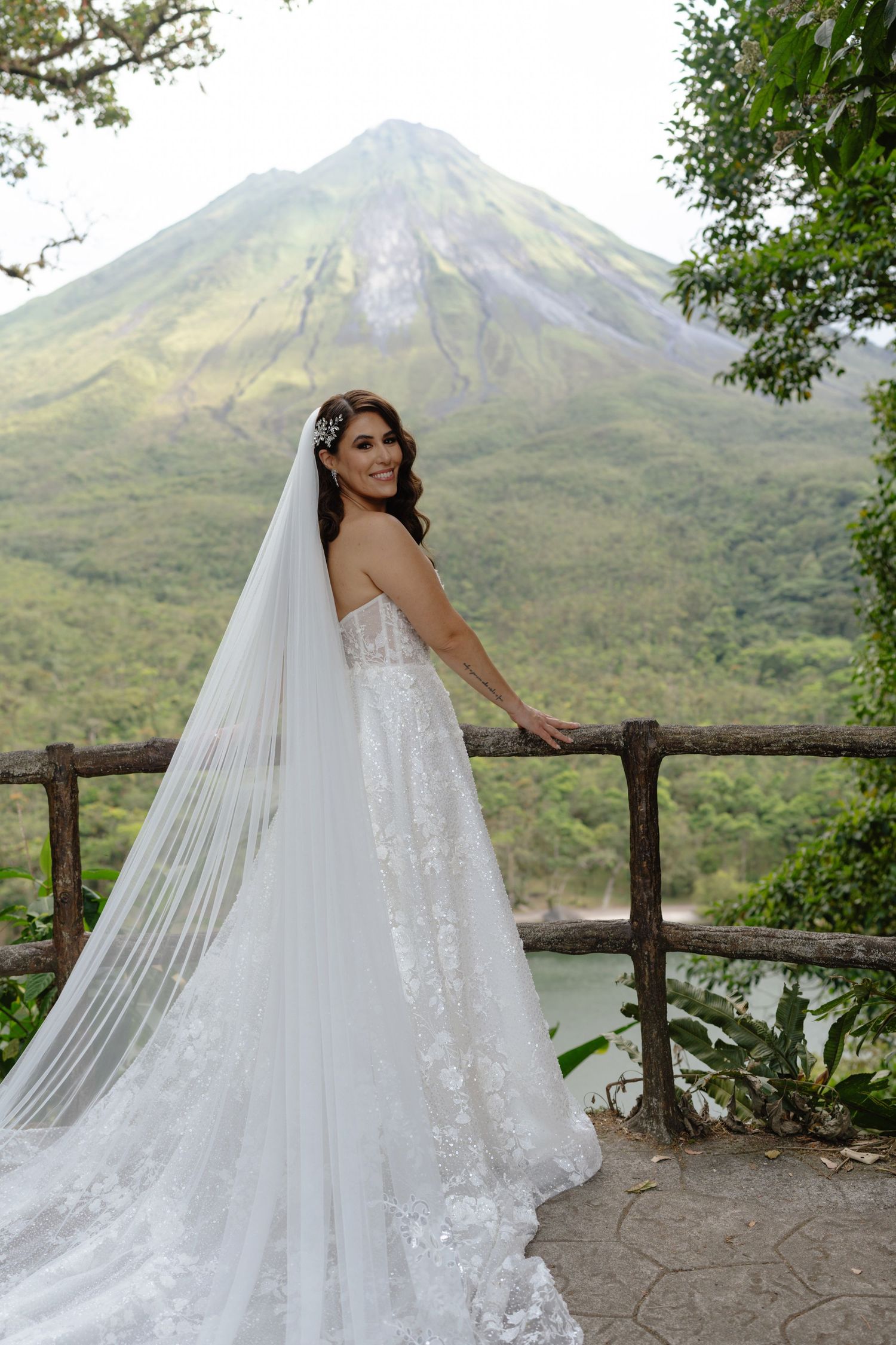 A wedding dress with flowing veil photographed against a scenic mountain backdrop with wooden railing.