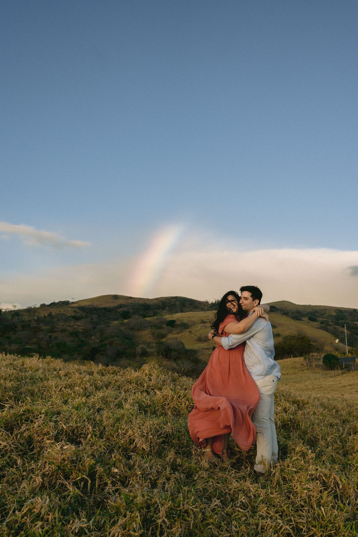A couple dances together in a golden field at sunset with rolling hills in the background.