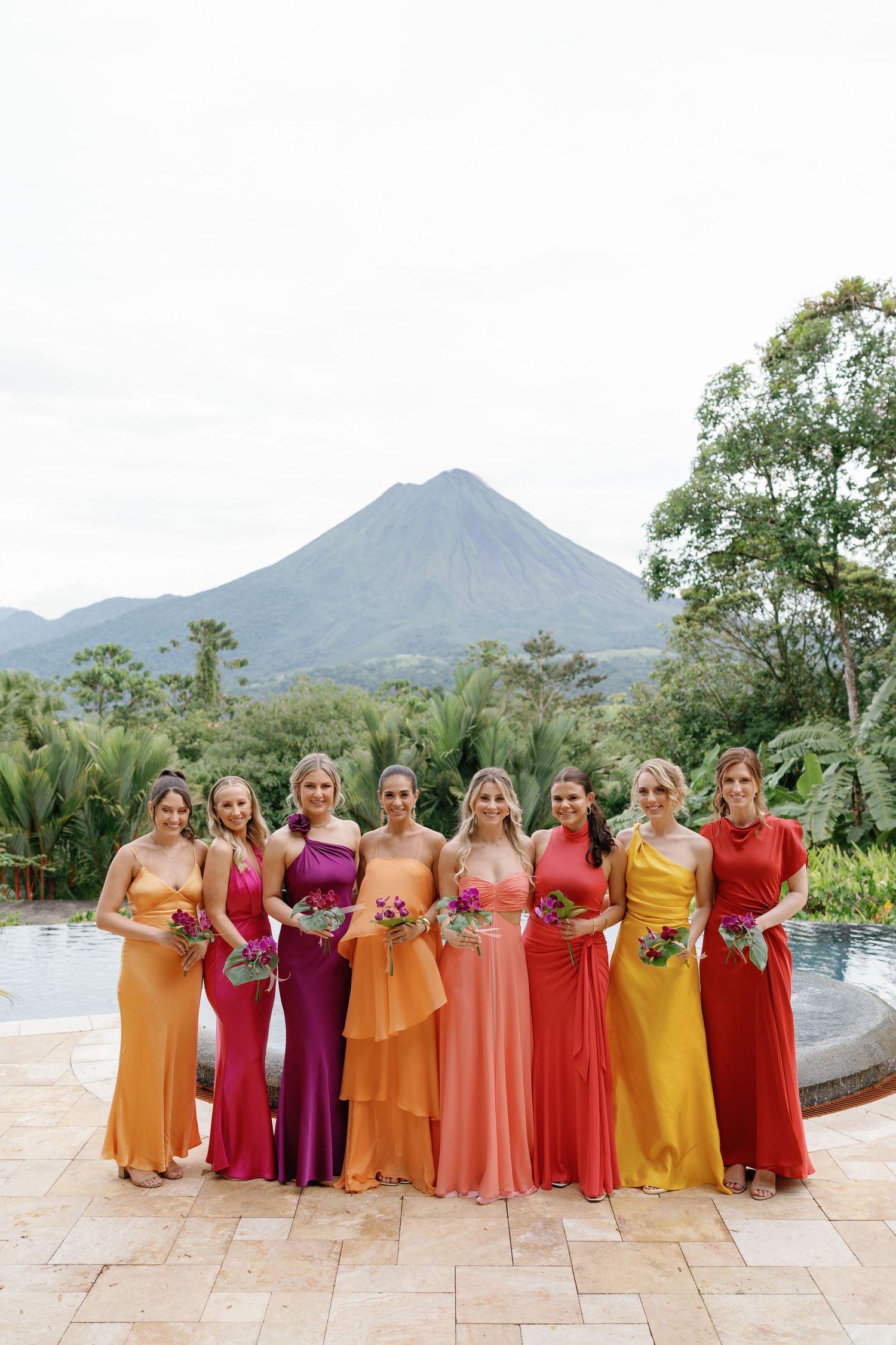 Bridesmaids in sunset-colored dresses pose together with mountain backdrop.