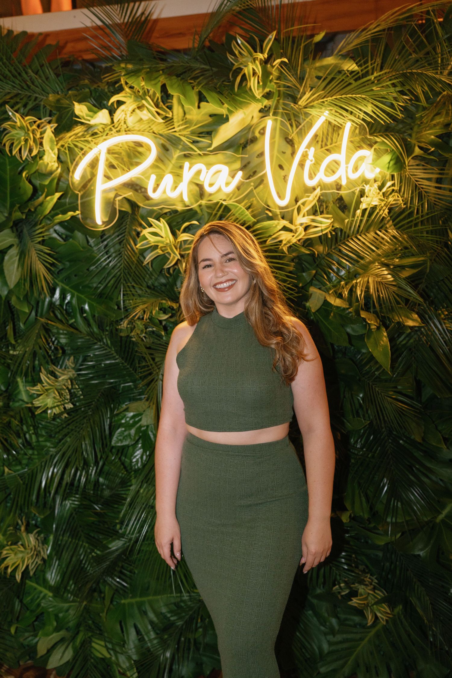 Person in olive green outfit poses against tropical leaf wall with neon sign.