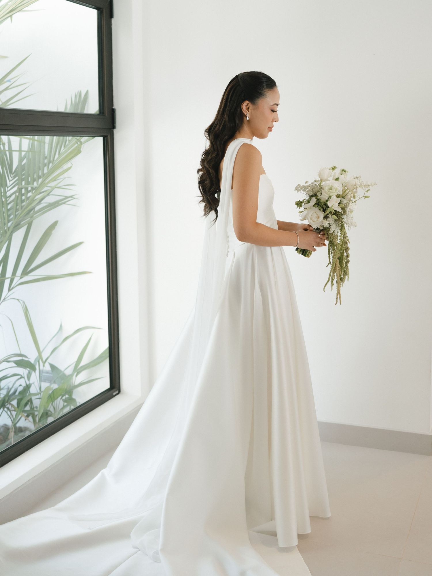 Bride in an elegant white wedding dress holds a bouquet while standing in a bright modern room with large windows.
