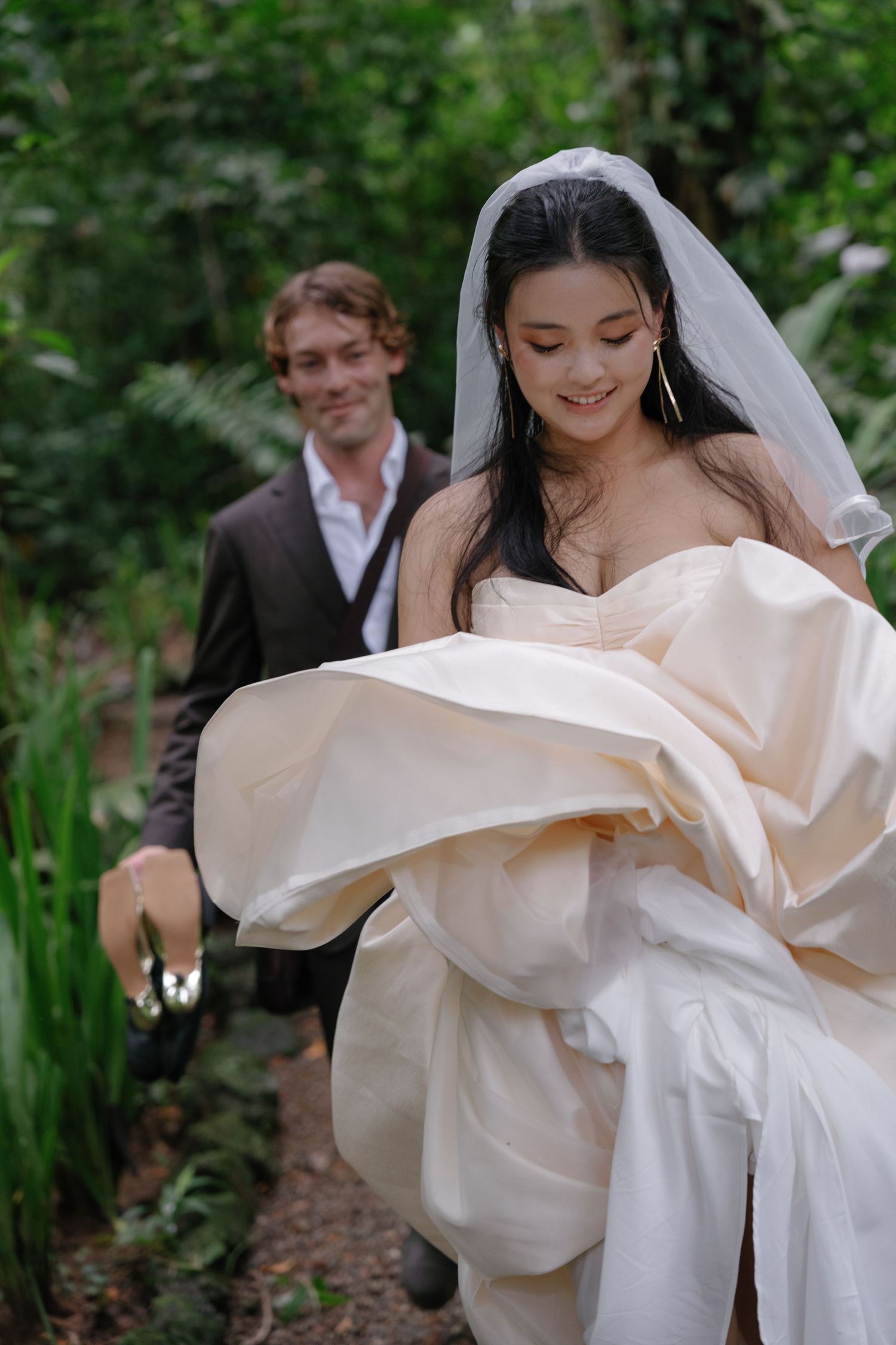 A bride in a flowing white wedding dress twirling outdoors among lush greenery.