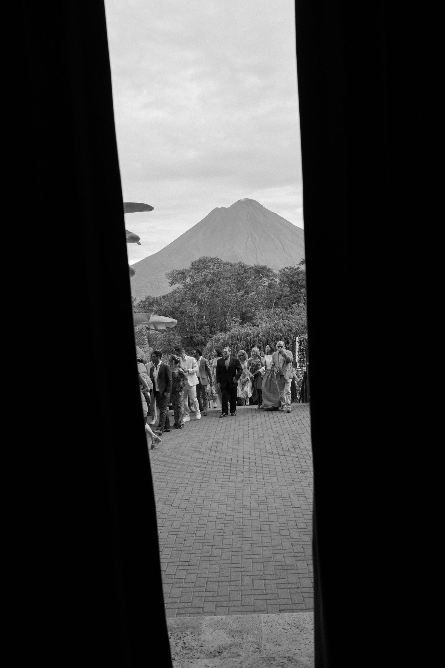 A crowd gathers on a brick path with a mountain peak visible in the distance through a framed view.