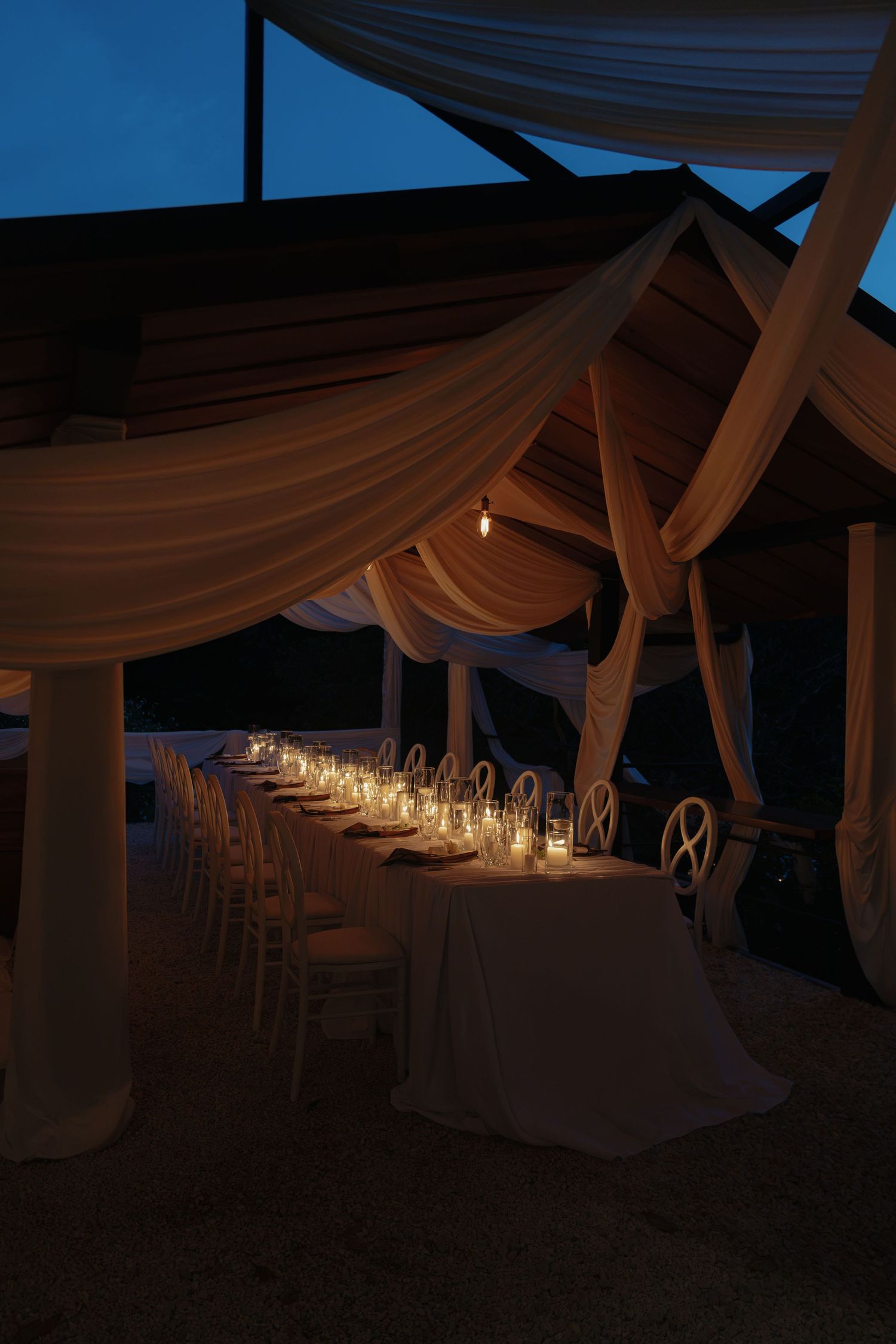 Elegant outdoor wedding reception table set with candles under draped white fabric at dusk.
