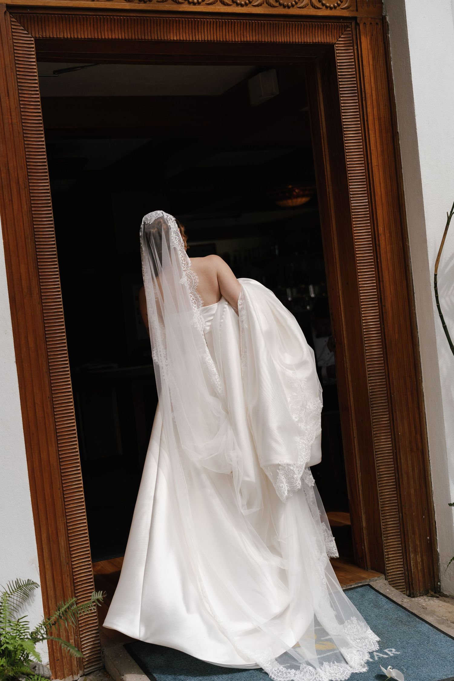 Bride in white wedding gown and veil entering through ornate wooden doorway, viewed from behind.