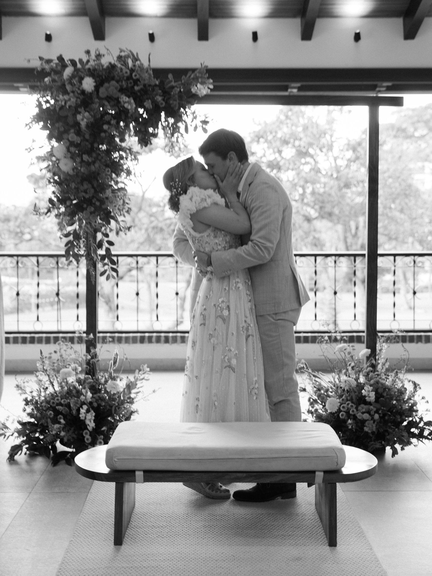 A romantic couple in formal attire shares a tender moment on a bench in a covered outdoor setting with floral decorations.