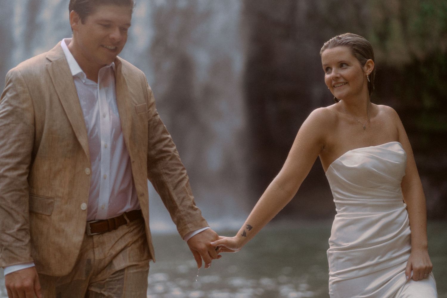 A couple in wedding attire shares a joyful moment by a waterfall.